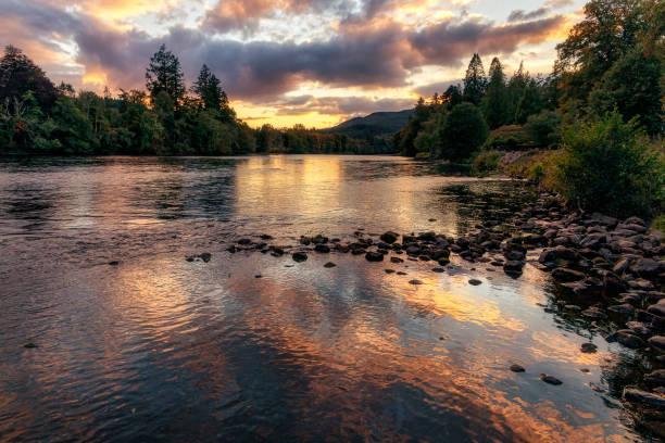 A calm river reflects a colorful sunset sky with clouds, surrounded by trees and distant hills.