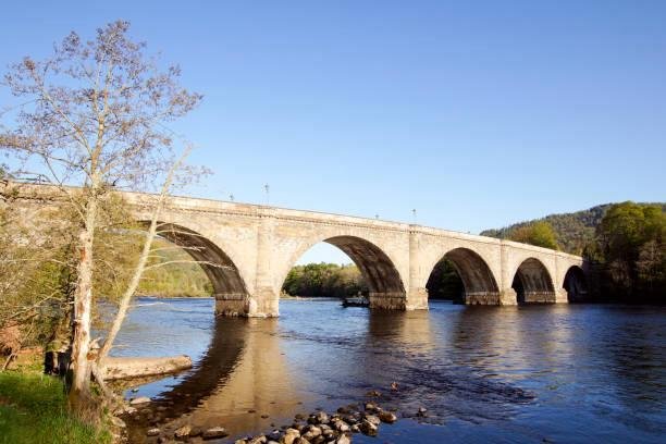 A stone bridge with multiple arches crossing over a river with trees on both sides and a clear blue sky.