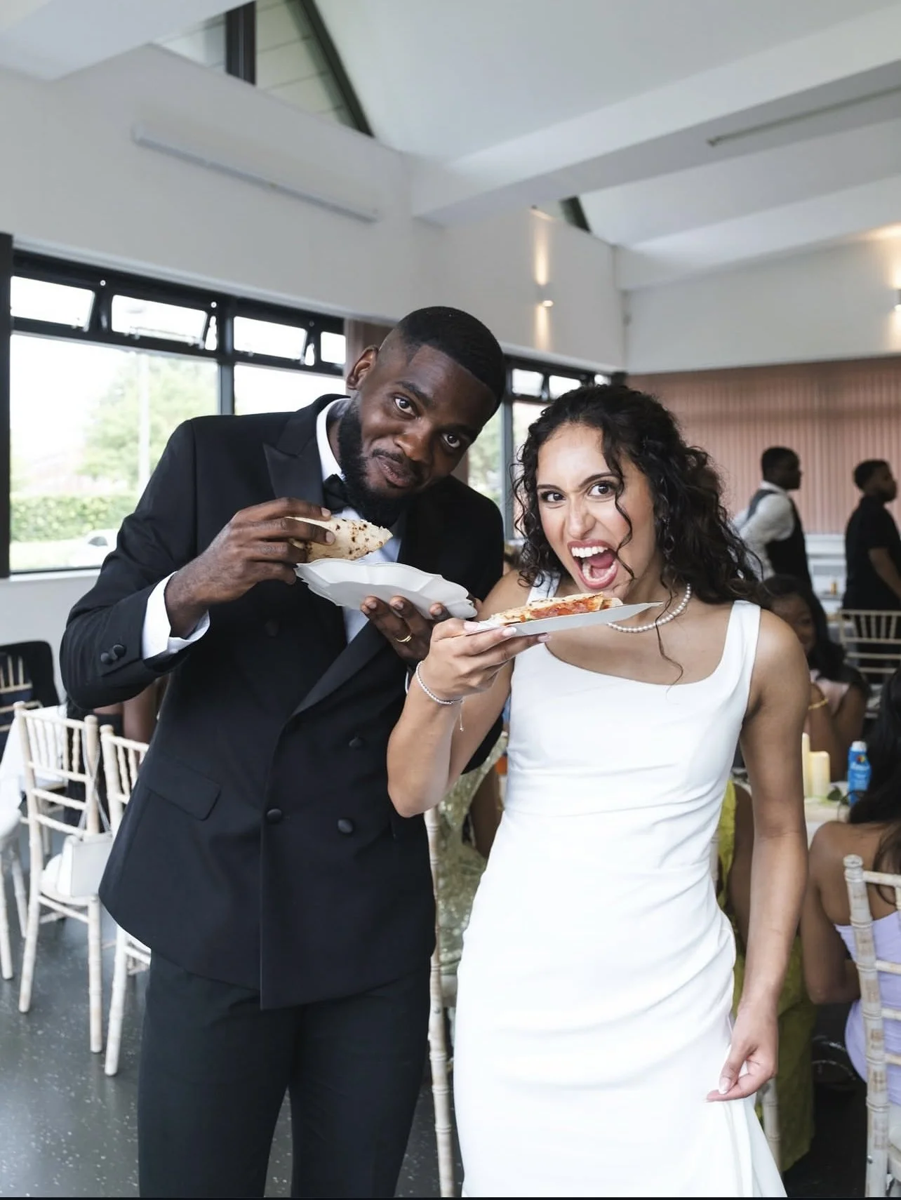 A happy woman in a white dress and a man in a black tuxedo are holding plates with slices of pizza at a celebration or wedding reception.
