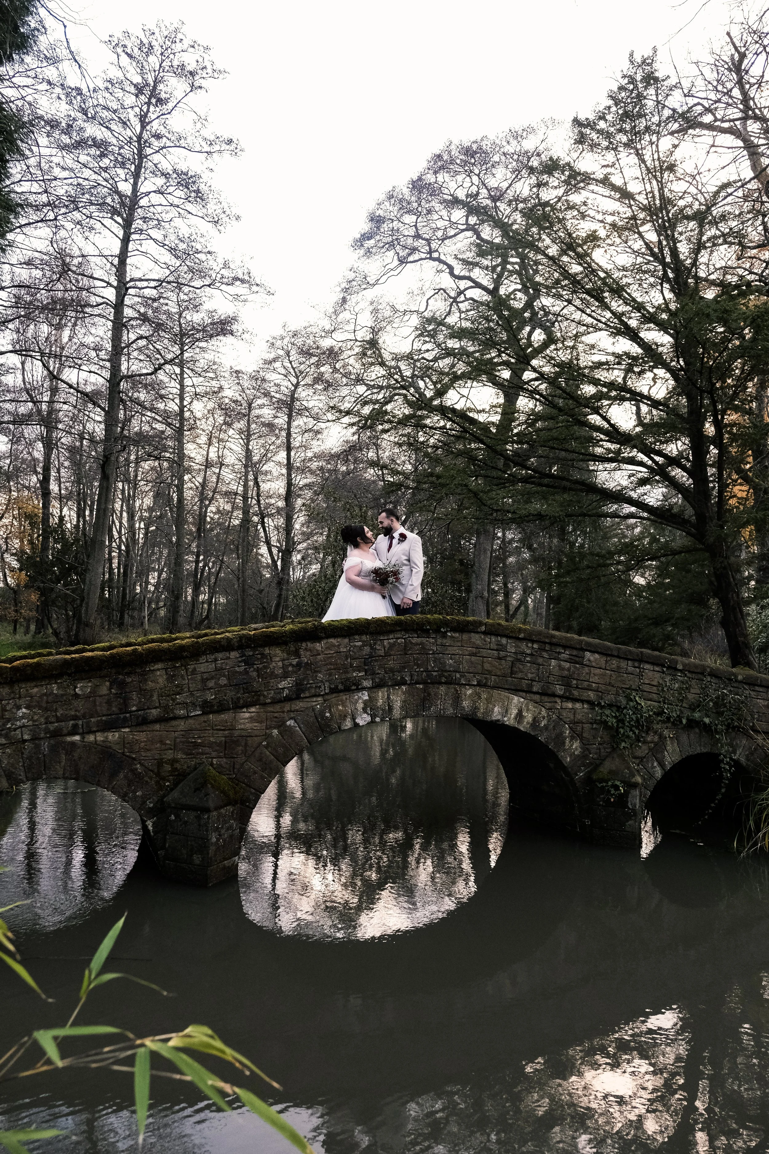 A bride and groom standing close together on a stone bridge over a river in a wooded park during dusk.