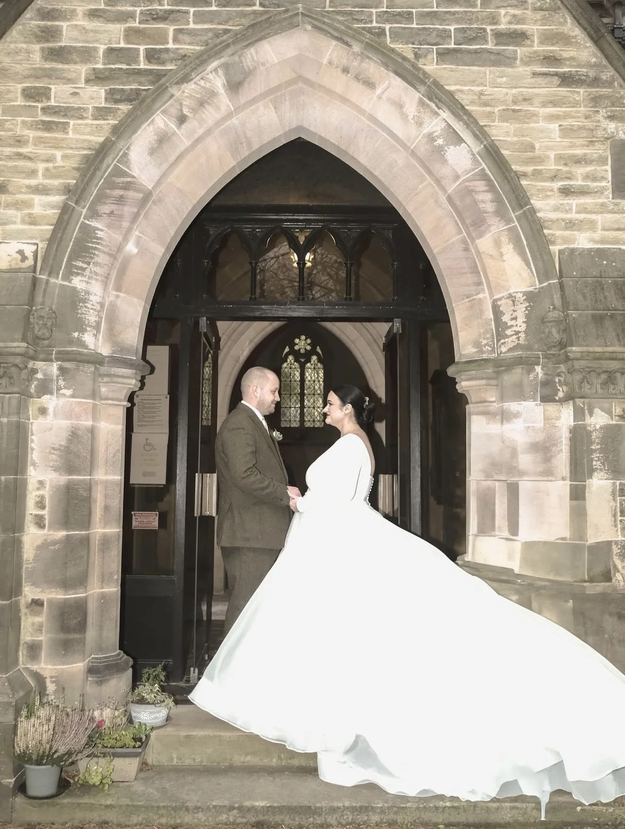 Bride and groom holding hands and facing each other in front of a church entrance, with the bride wearing a long white wedding gown and the groom in a brown suit, surrounded by stone architecture and stained glass windows.