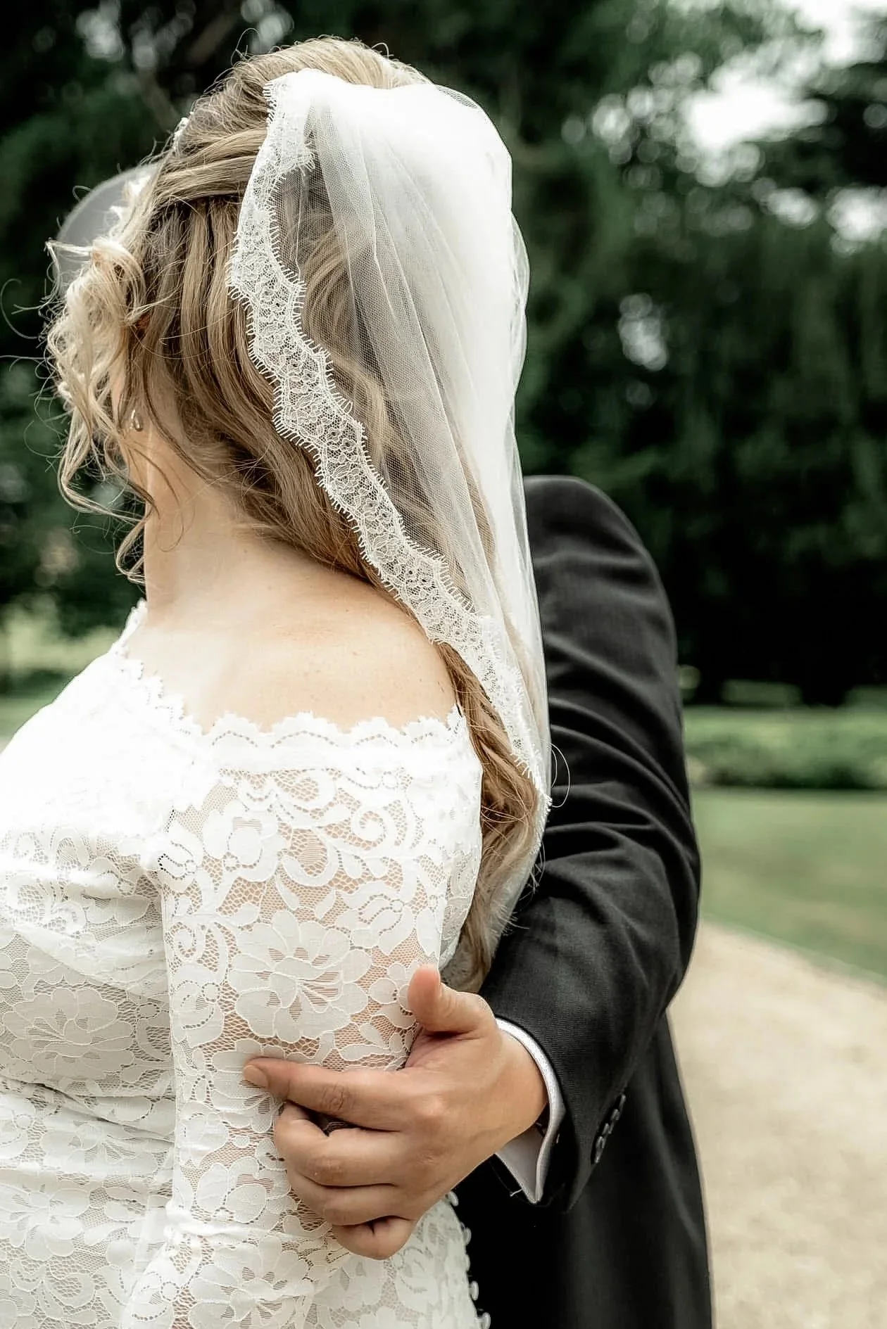 Close-up of a bride in a white lace wedding dress with a veil, being held by a groom in a black suit outdoors.