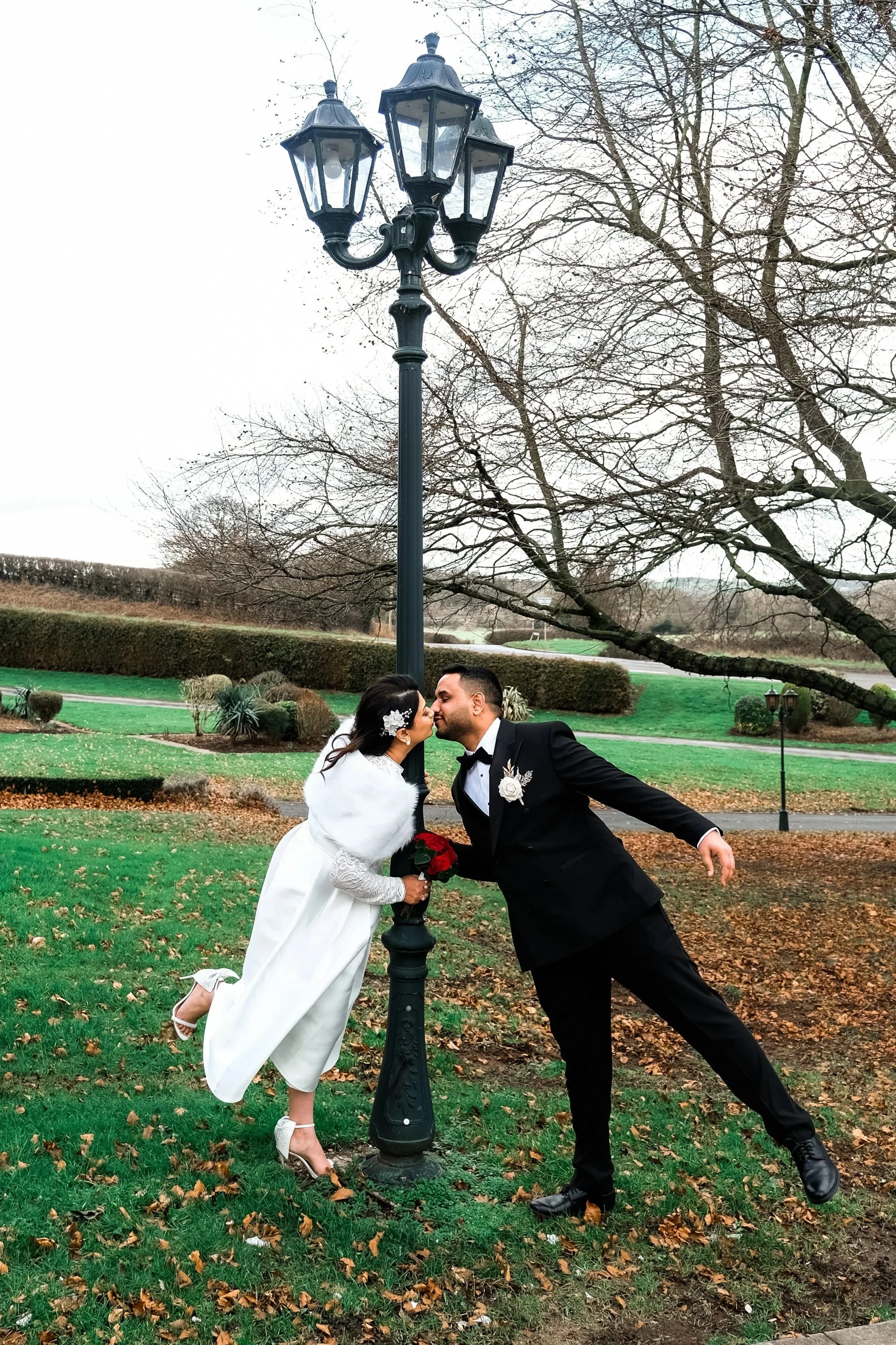 A bride and groom leaning in to kiss under a black street lamp in a park with green grass and leafless trees.