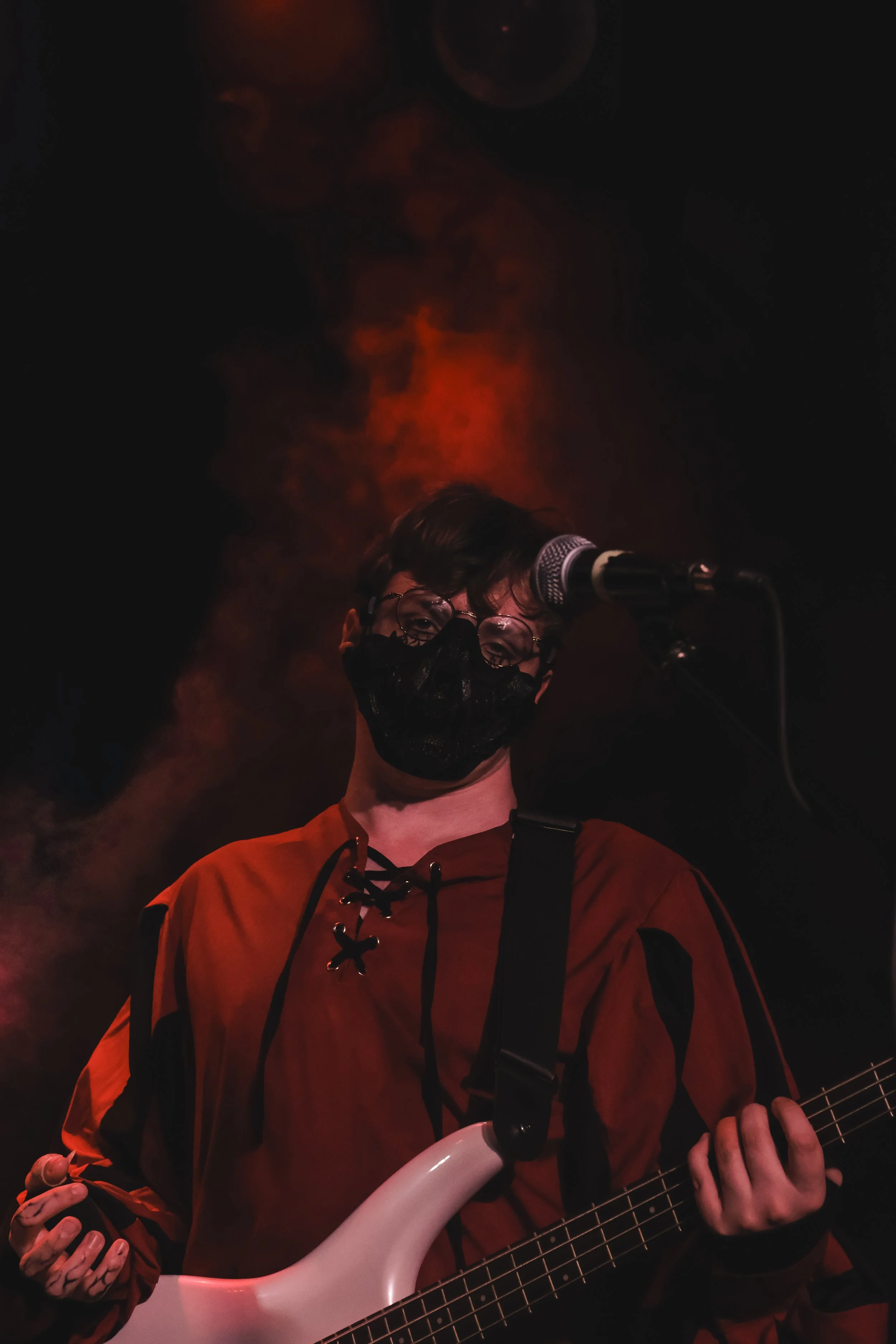 A person wearing glasses and a black face mask, playing a white electric guitar on stage with dark and orange lighting.