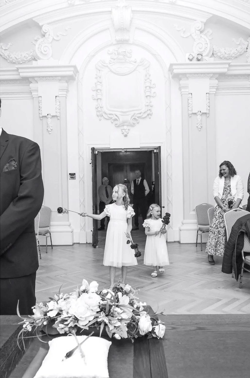 Two young girls in white dresses holding roses walk down an aisle during a wedding ceremony in a decorated hall. Flowers rest on a table in the foreground.