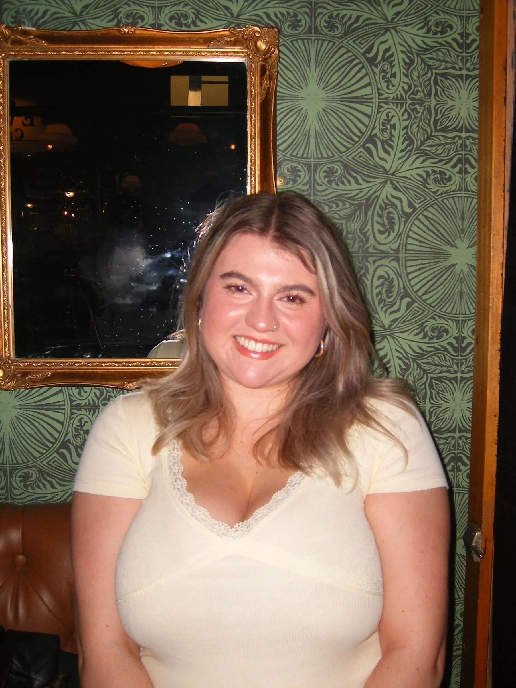 A woman with shoulder-length light brown hair smiling, wearing a light yellow top, standing in front of a green patterned wall and a gold-framed mirror.