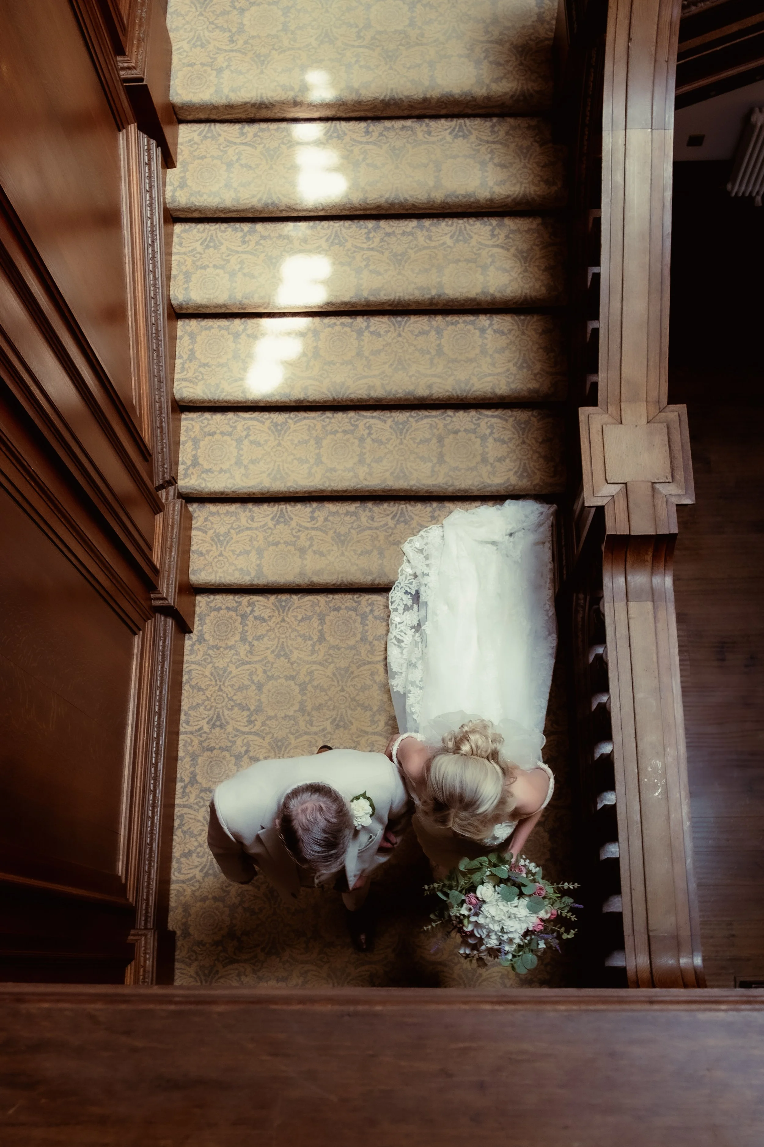 Second from bottom view of a bride and her father standing on a carpeted staircase, taken from above.