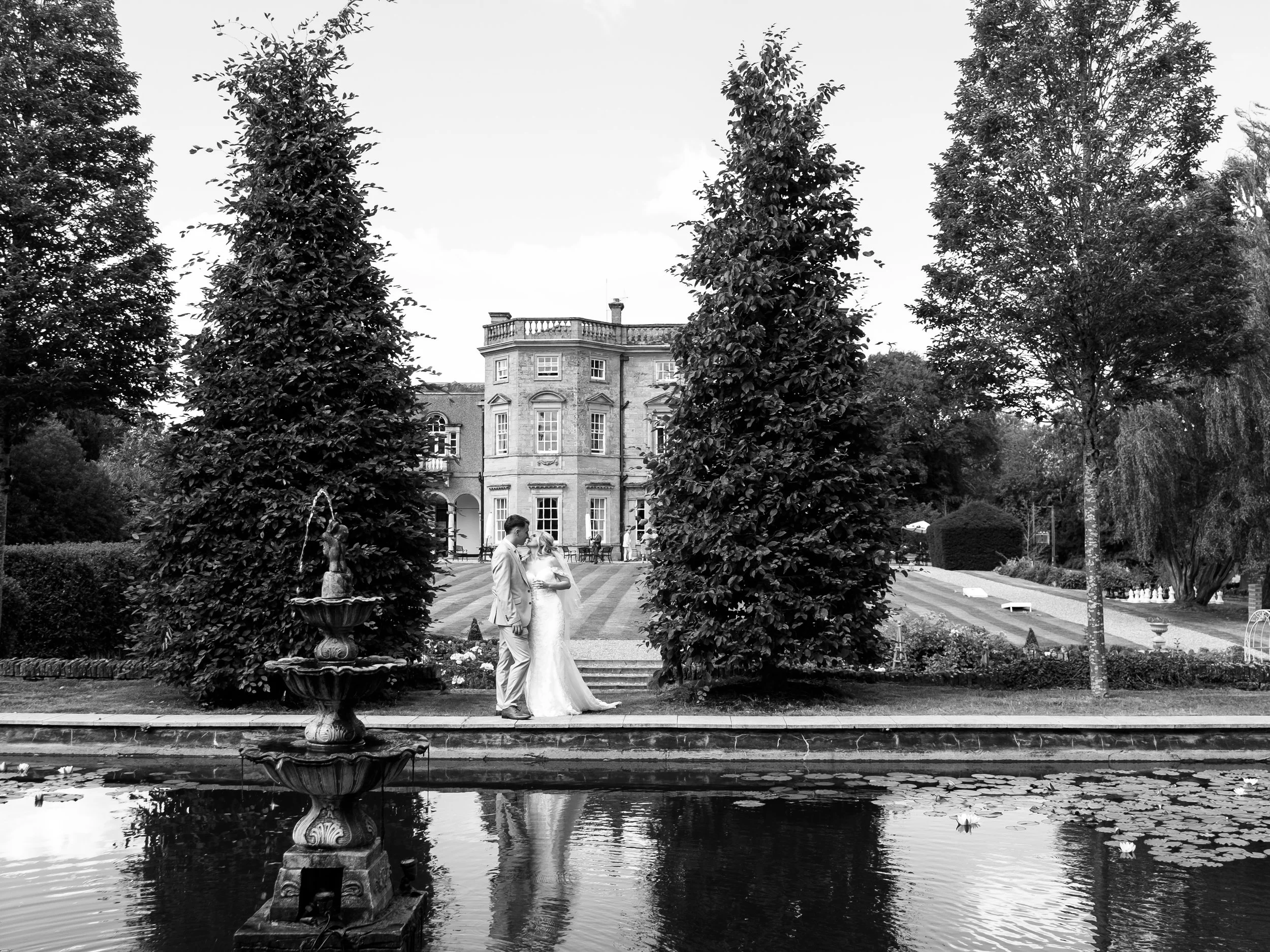 A black and white photo of a wedding couple standing beside each other in a garden with a fountain in the foreground, trees, and a large historic building in the background.