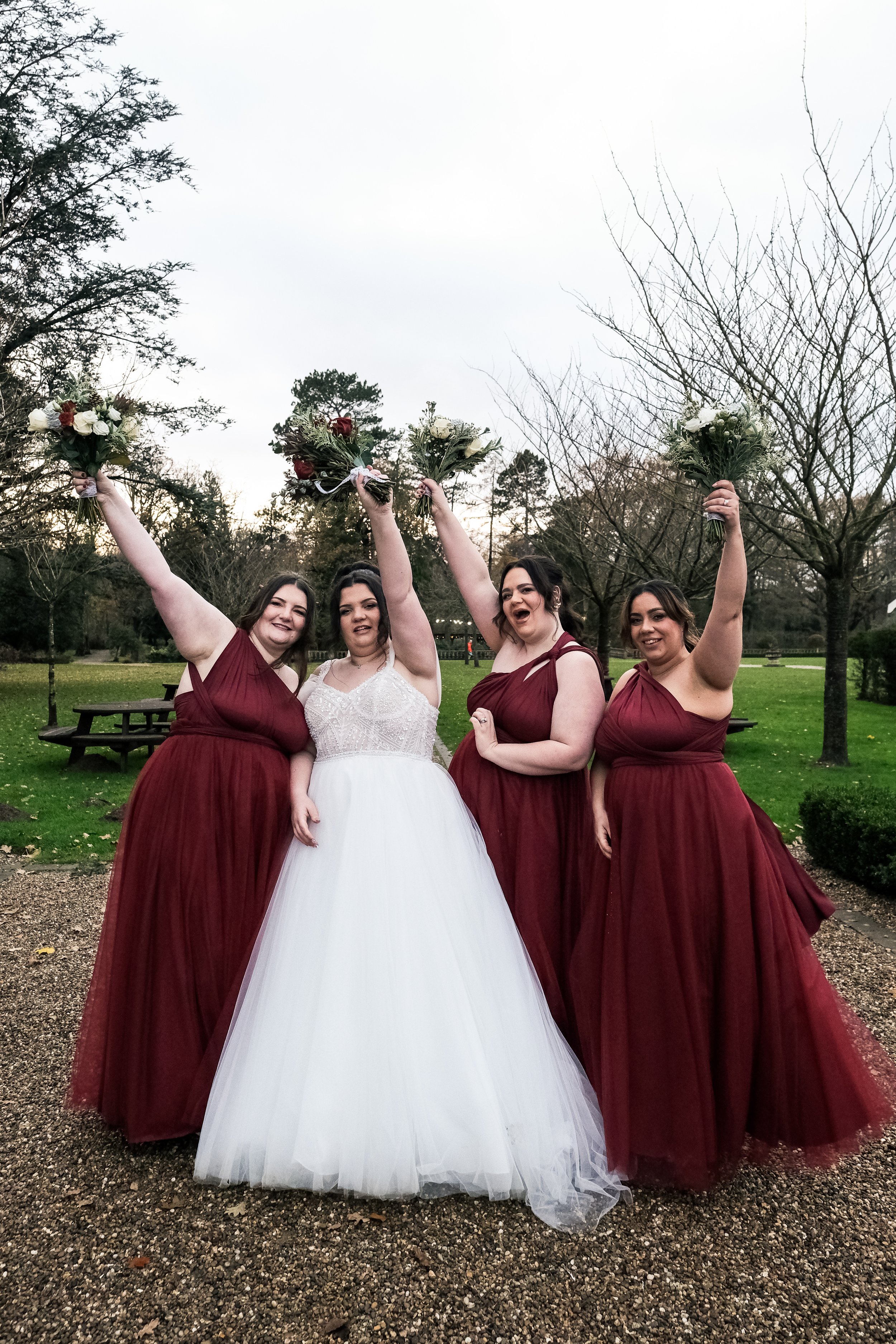 A bride and three bridesmaids celebrating outdoors, all holding bouquets and smiling. The bride is in a white gown, and the bridesmaids wear matching maroon dresses. The setting is a park with leafless trees and green grass.
