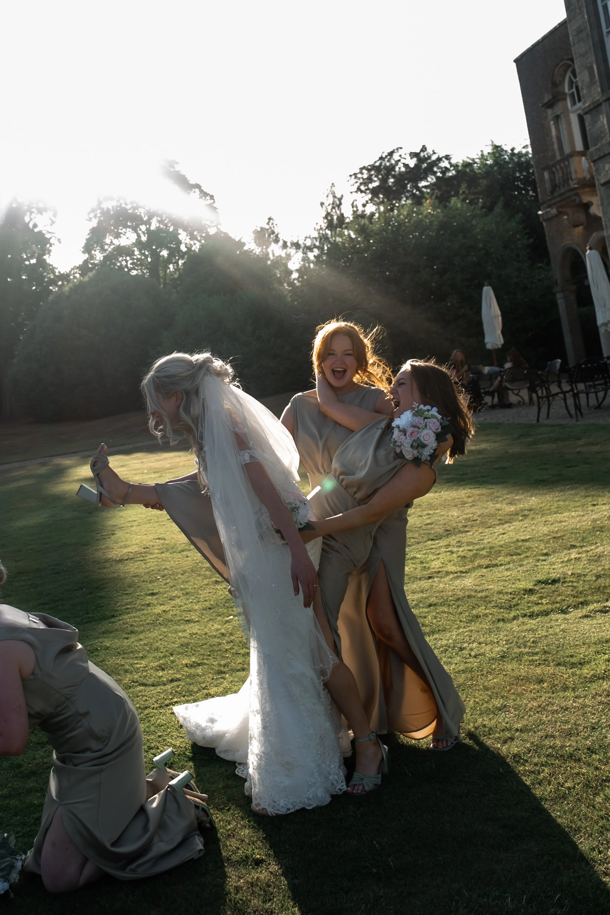 Three women, including a bride in a white wedding dress, having fun outdoors on a sunny day; two women are lifting the bride, smiling and laughing, in a garden or park area with trees, chairs, and an elegant building in the background.