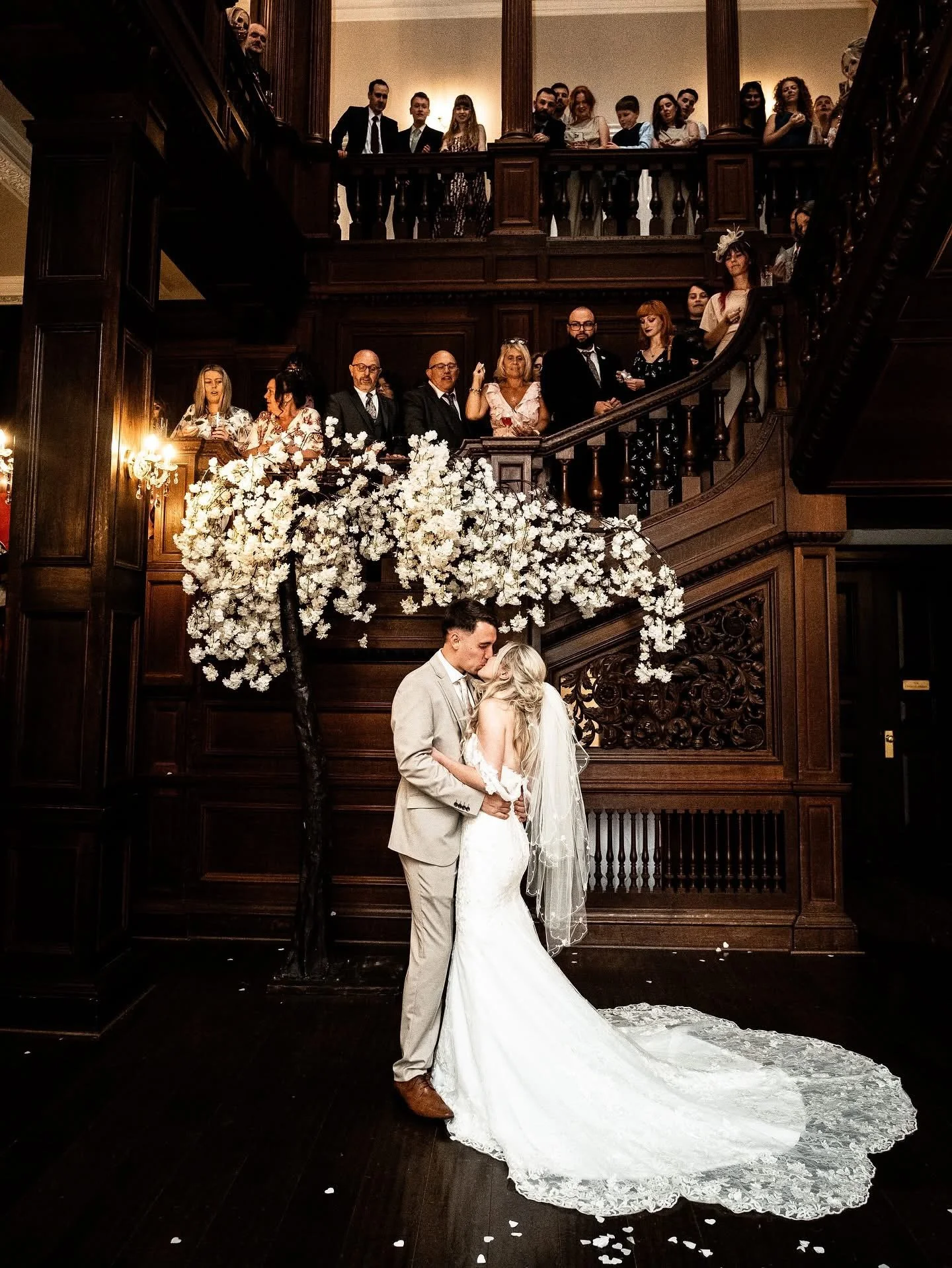 A newlywed couple sharing a kiss under a floral arch at their wedding ceremony, surrounded by guests observing from multiple levels of a dark wood staircase in an elegant venue.