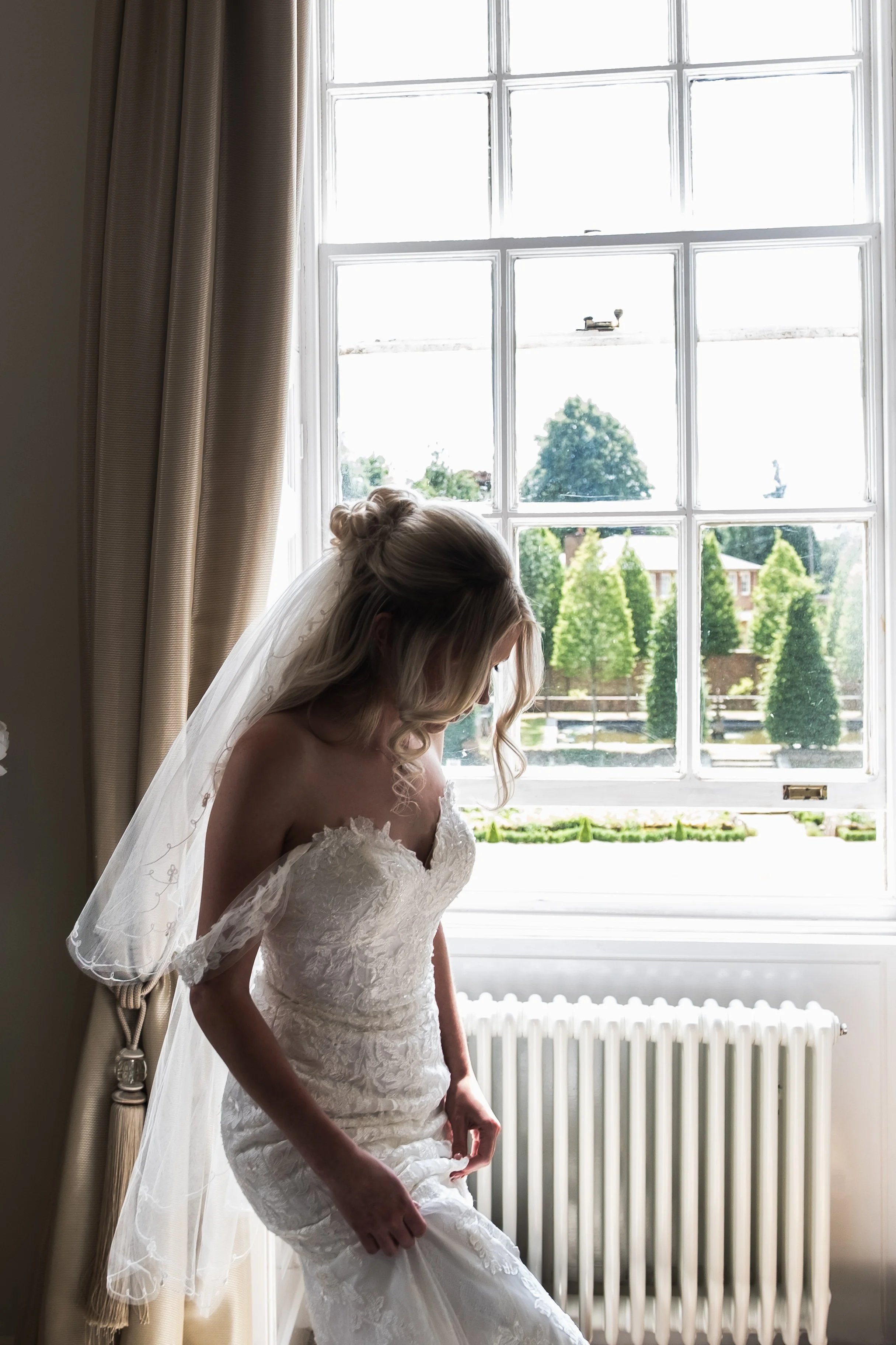 Bride in a white wedding gown standing by a window, adjusting her dress, with light streaming in and a view of trees outside.
