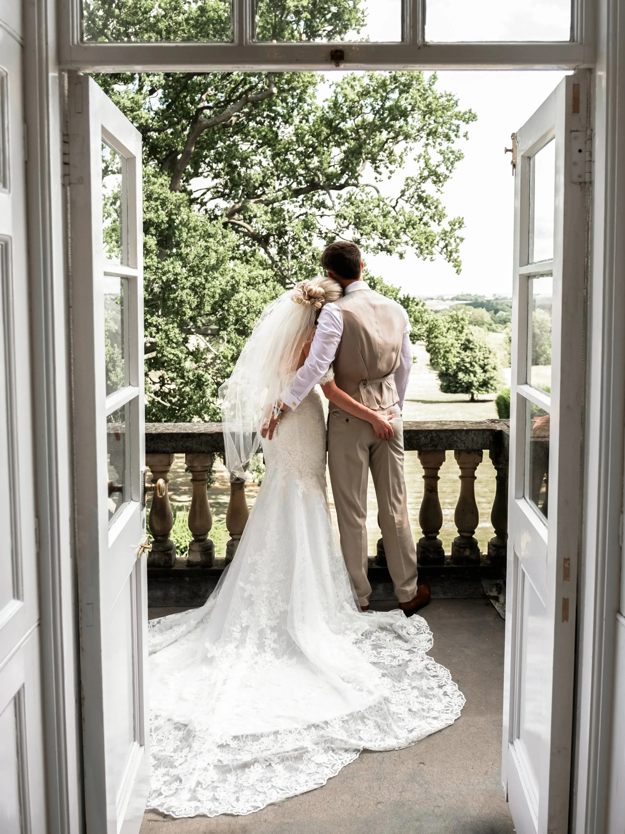 A bride and groom are standing together on a balcony, embracing and looking at a scenic landscape with trees.