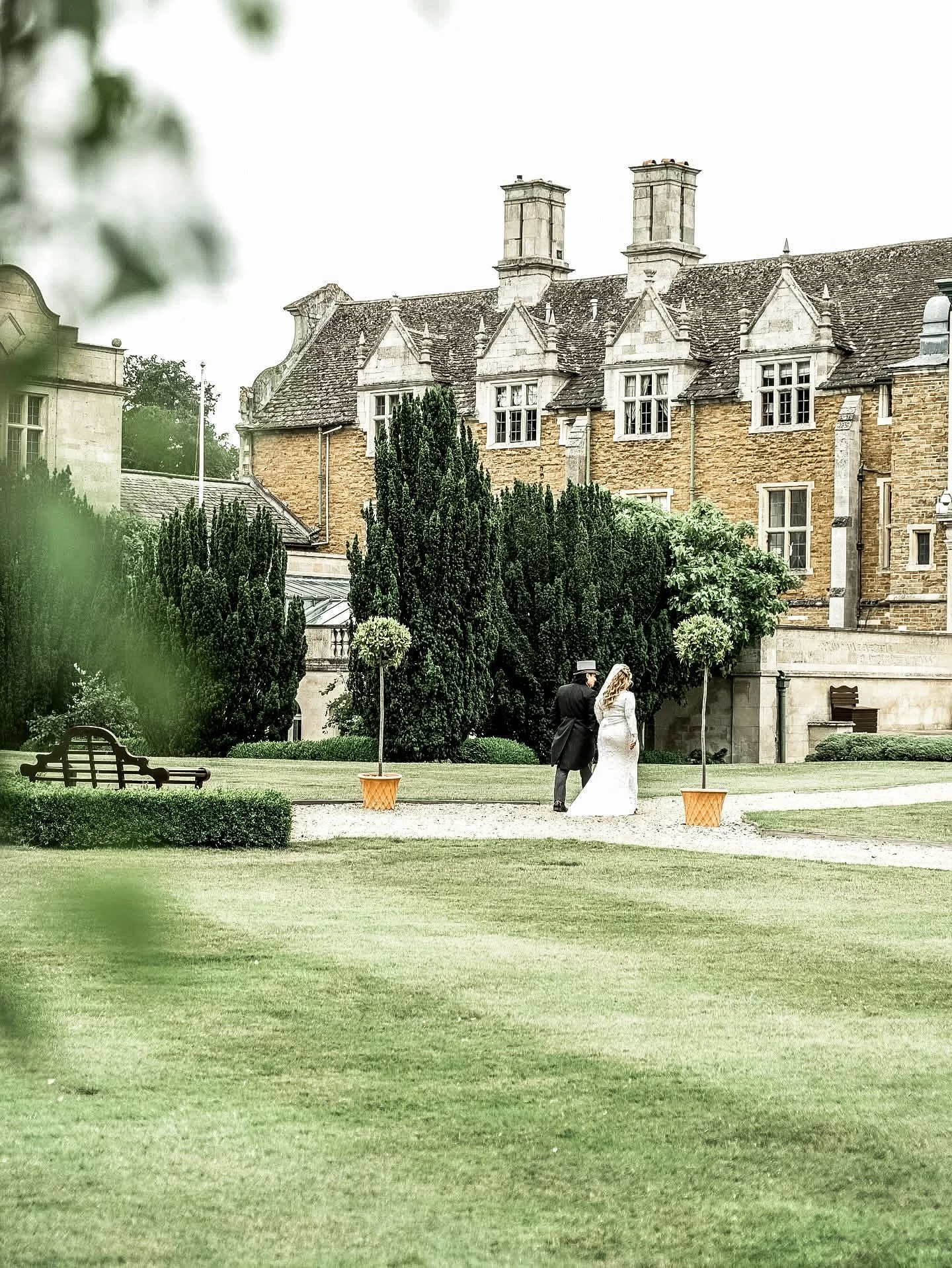 A bride and groom walking on a pathway in a garden with large trees and a historic building in the background.