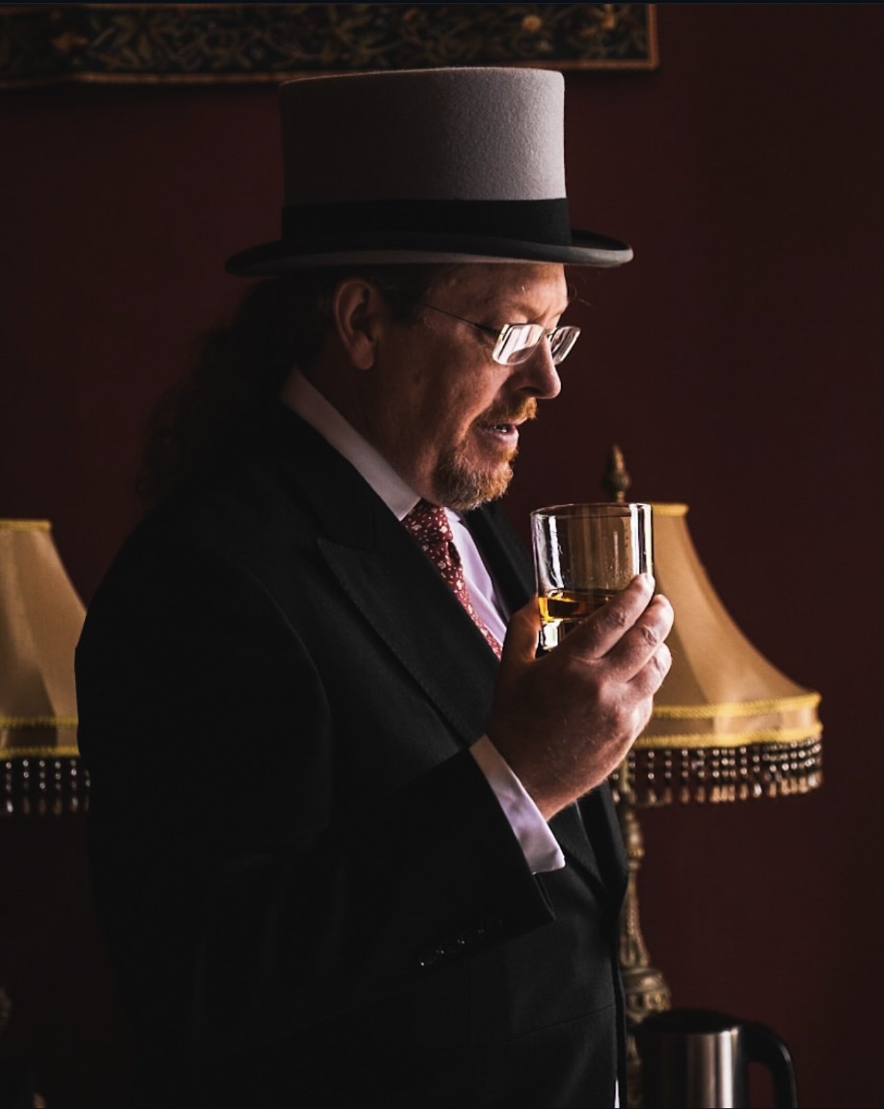 A man in a tuxedo and a tall gray hat, holding a glass of whiskey, stands in a dimly lit room with ornate lamps and dark red walls.