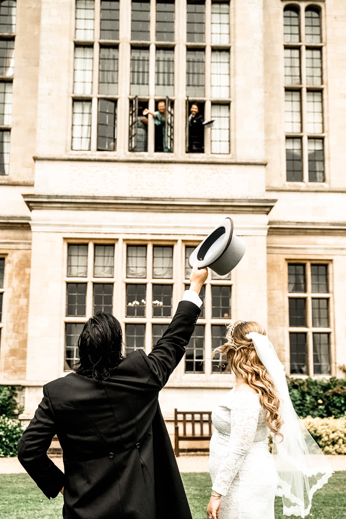A bride and groom are outside in front of a large building, with the groom raising a top hat in one hand as he looks at the bride. The bride is wearing a lace wedding dress and has a veil, while the groom is dressed in a black tuxedo. Two men are loo