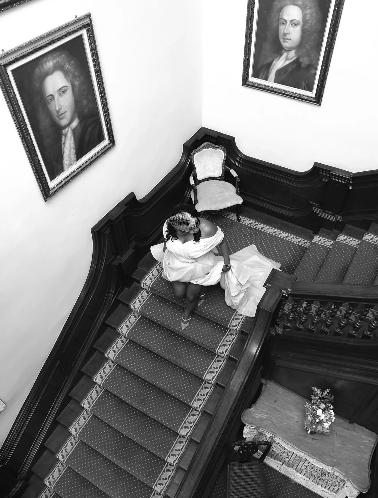 A woman in a wedding dress sitting on a staircase in a grand, ornate interior with large portrait paintings on the wall.