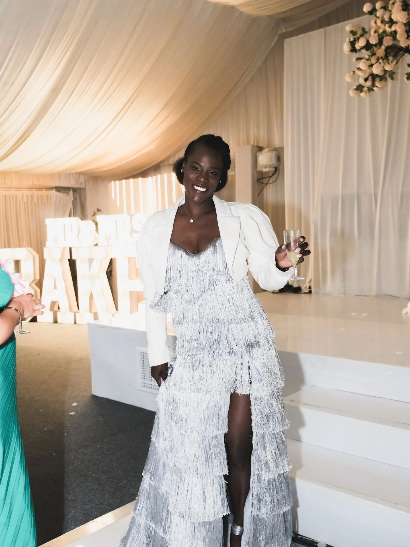 A woman in a glamorous silver evening dress with layered fringe detail and a white blazer, holding a glass of champagne, smiling at a celebration event.