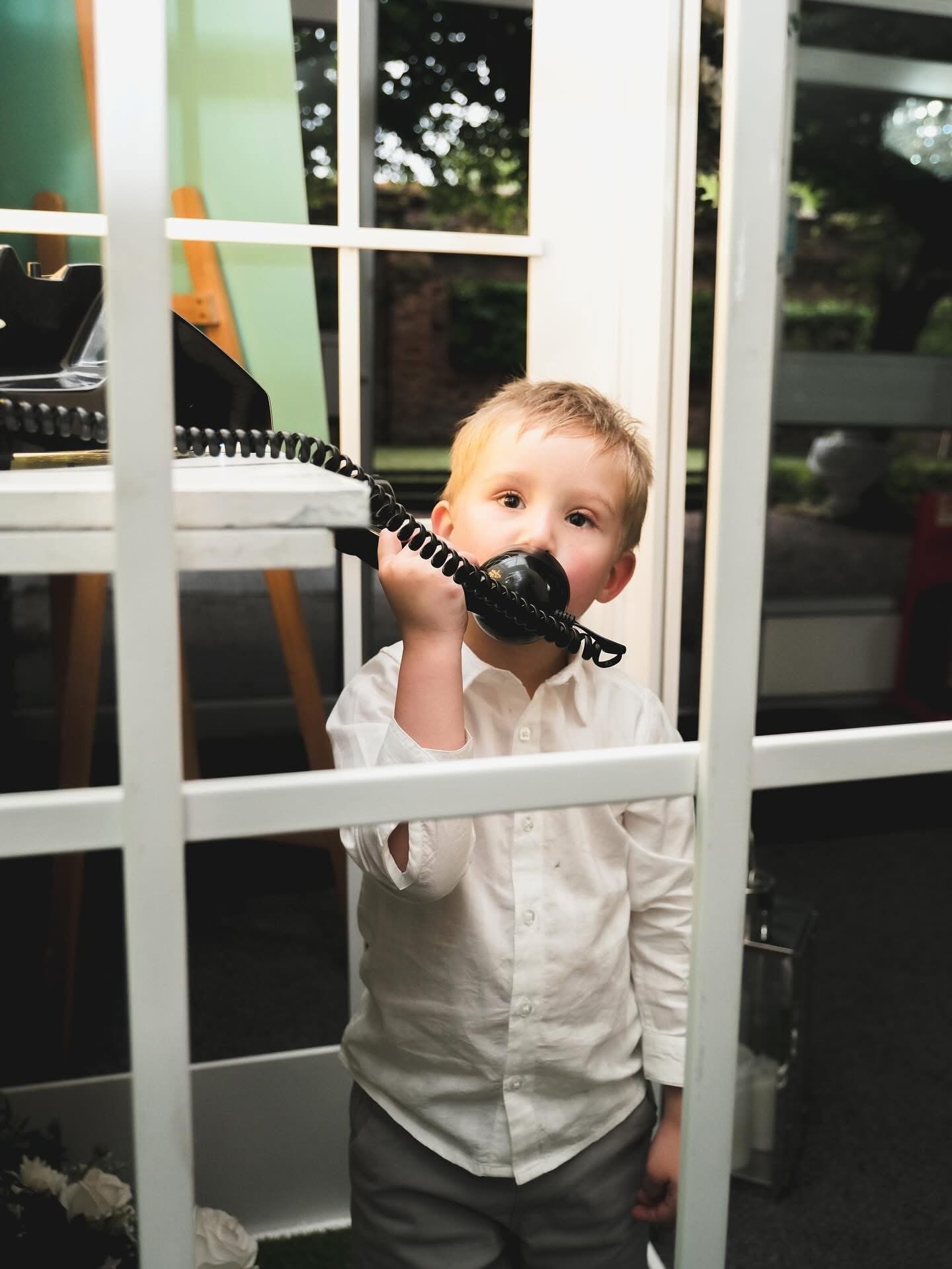 Young boy in white shirt holding a black rotary telephone, standing behind a white metal grid or fence, with an outdoor view through the window in the background.