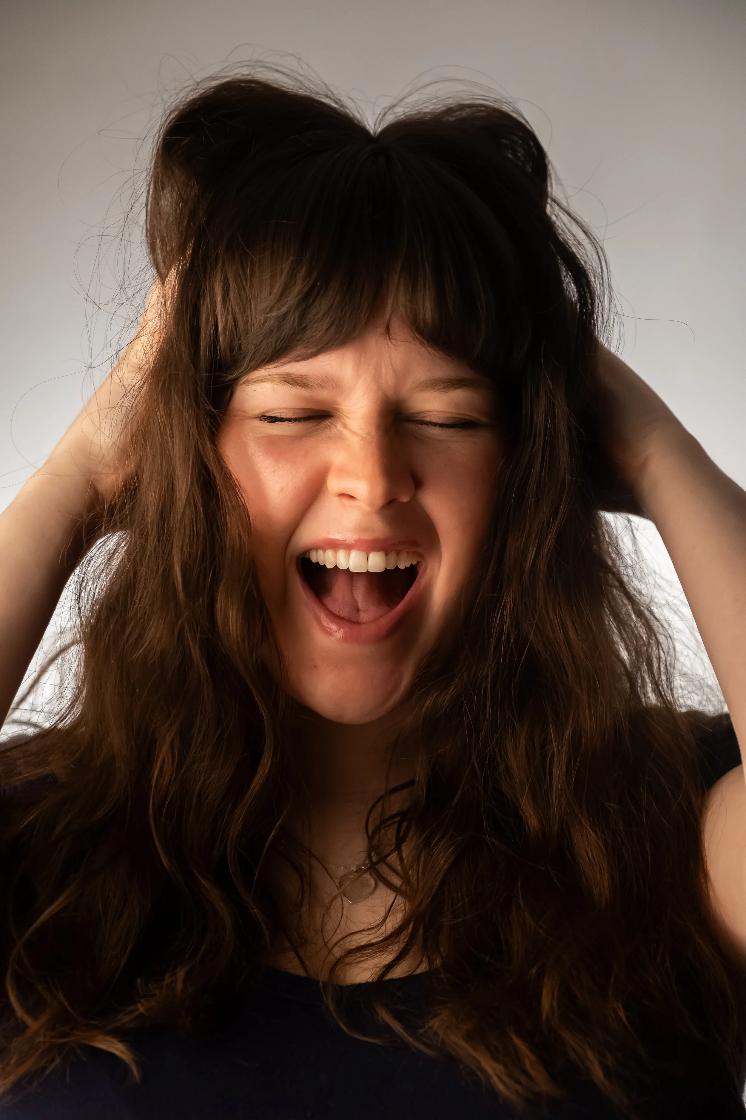 A woman with long wavy brown hair is holding her head with both hands, eyes closed, mouth wide open, appearing excited and confident.