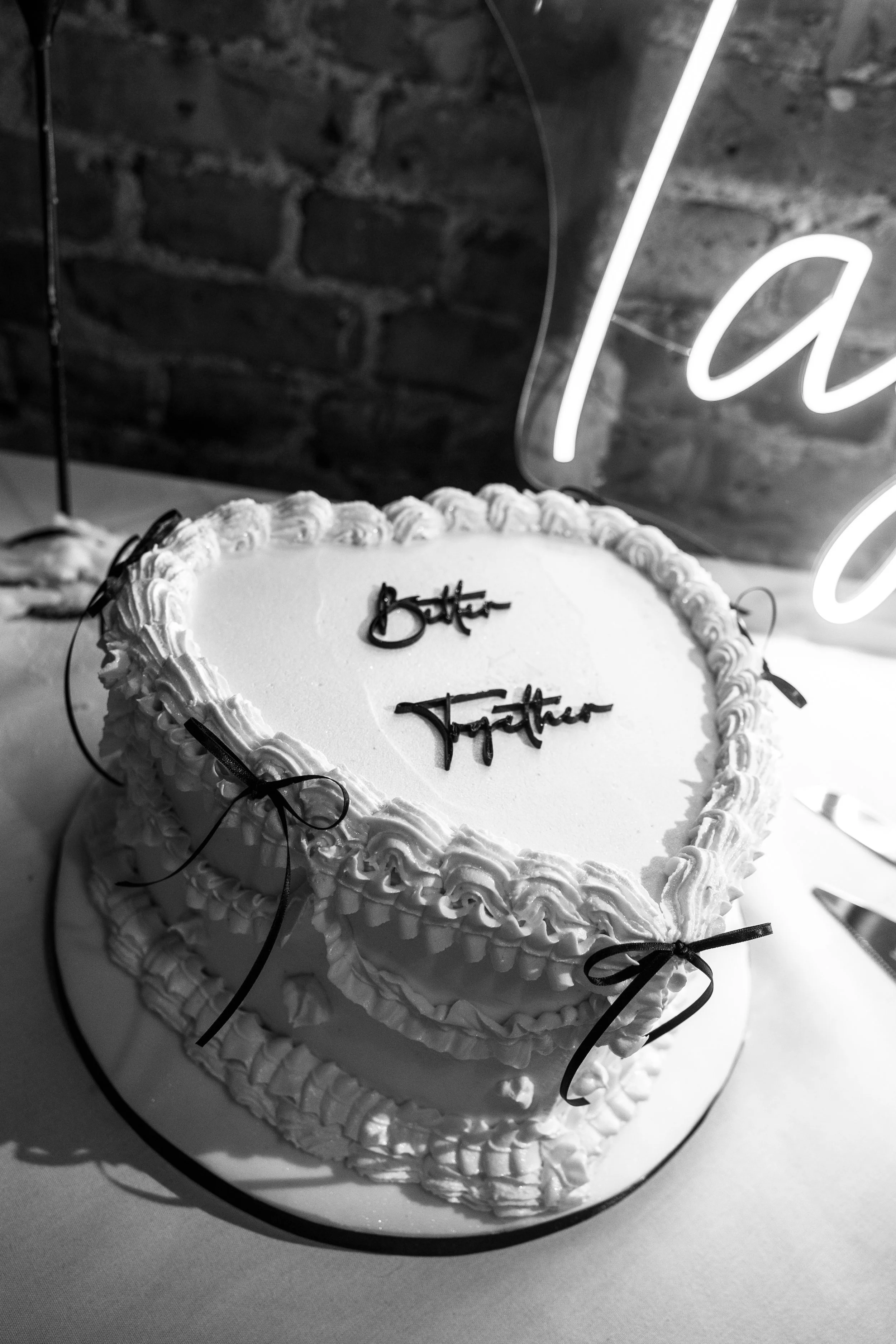 Heart-shaped wedding cake with white icing and black cursive writing that says 'Better' and 'Together', decorated with small bows and icing borders.