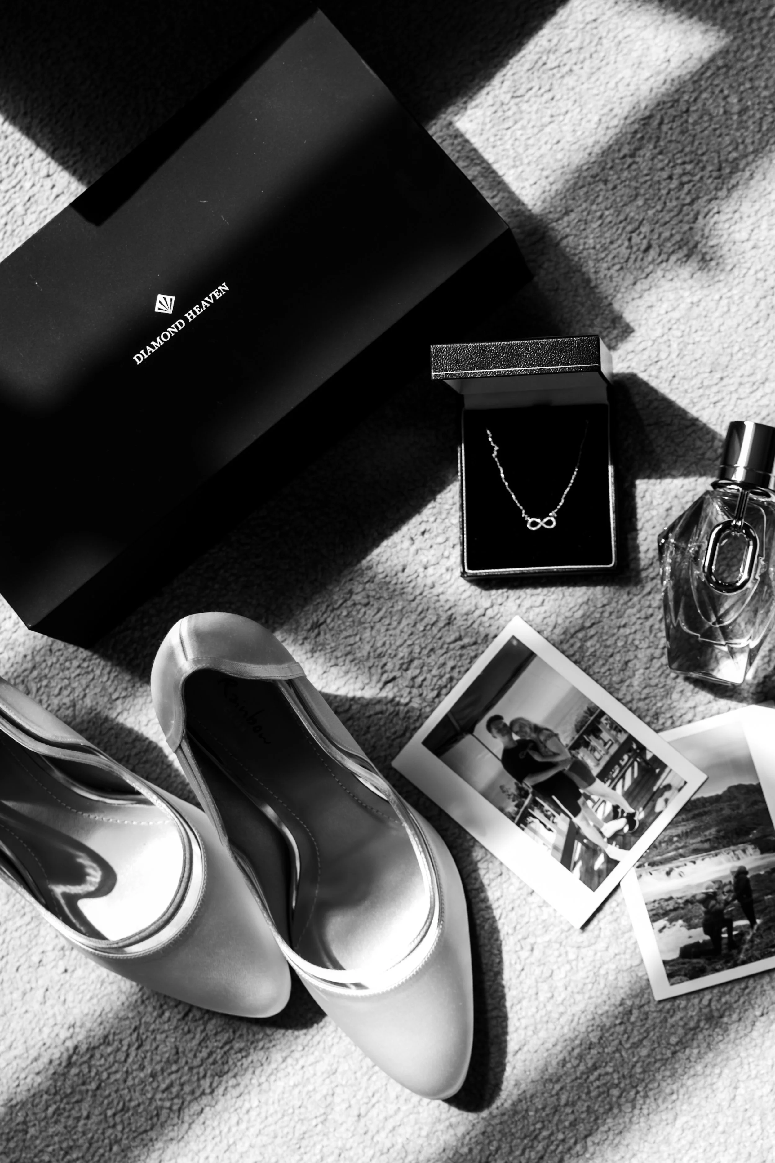 A black and white photo featuring a pair of shiny high-heeled shoes, a Diamond Heaven jewelry box with a necklace, a small perfume bottle, two photographs, and an open jewelry box with a necklace inside, all on a carpeted floor with light and shadow.