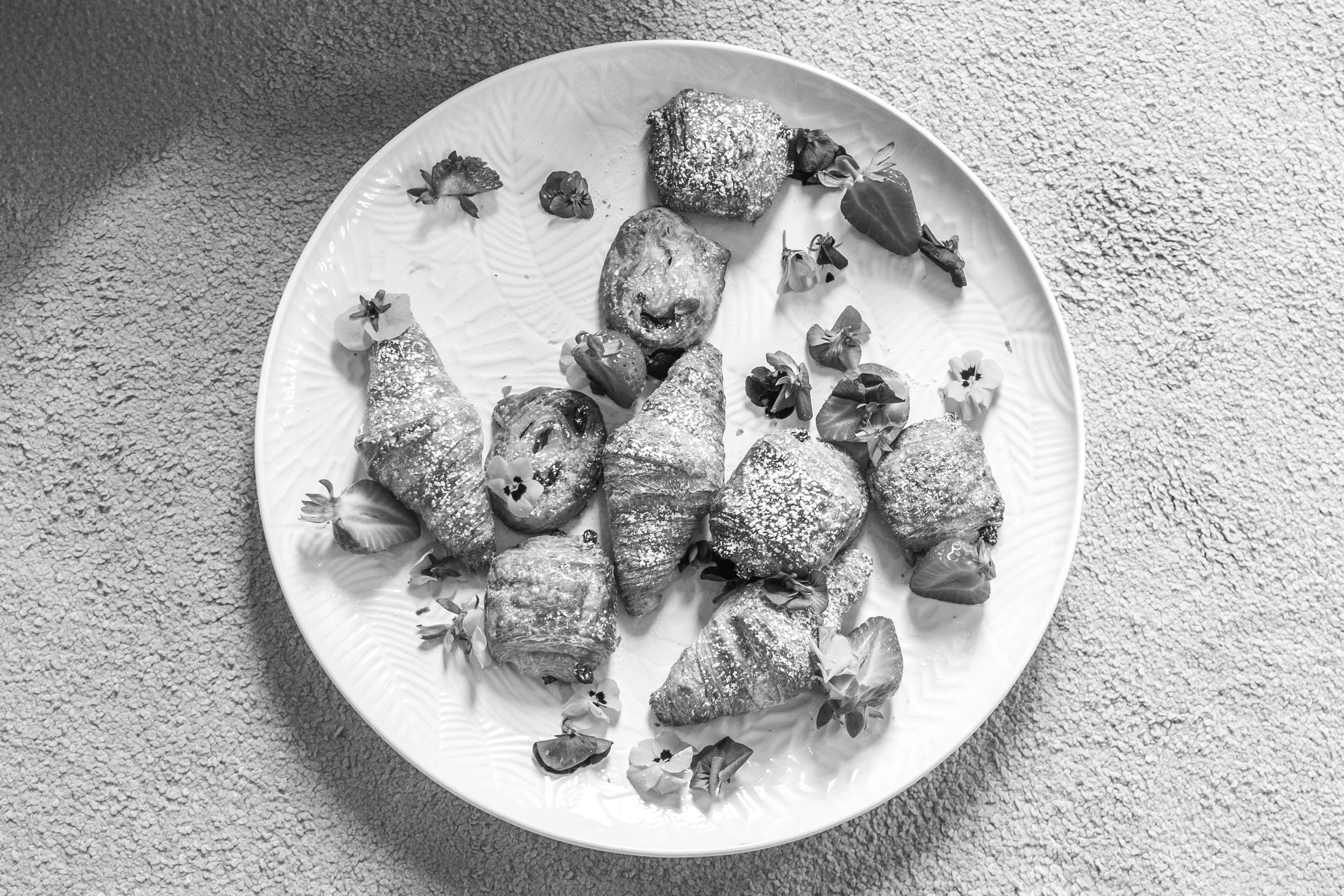 A white plate with assorted pastries dusted with powdered sugar, garnished with edible flowers, set on a textured surface.