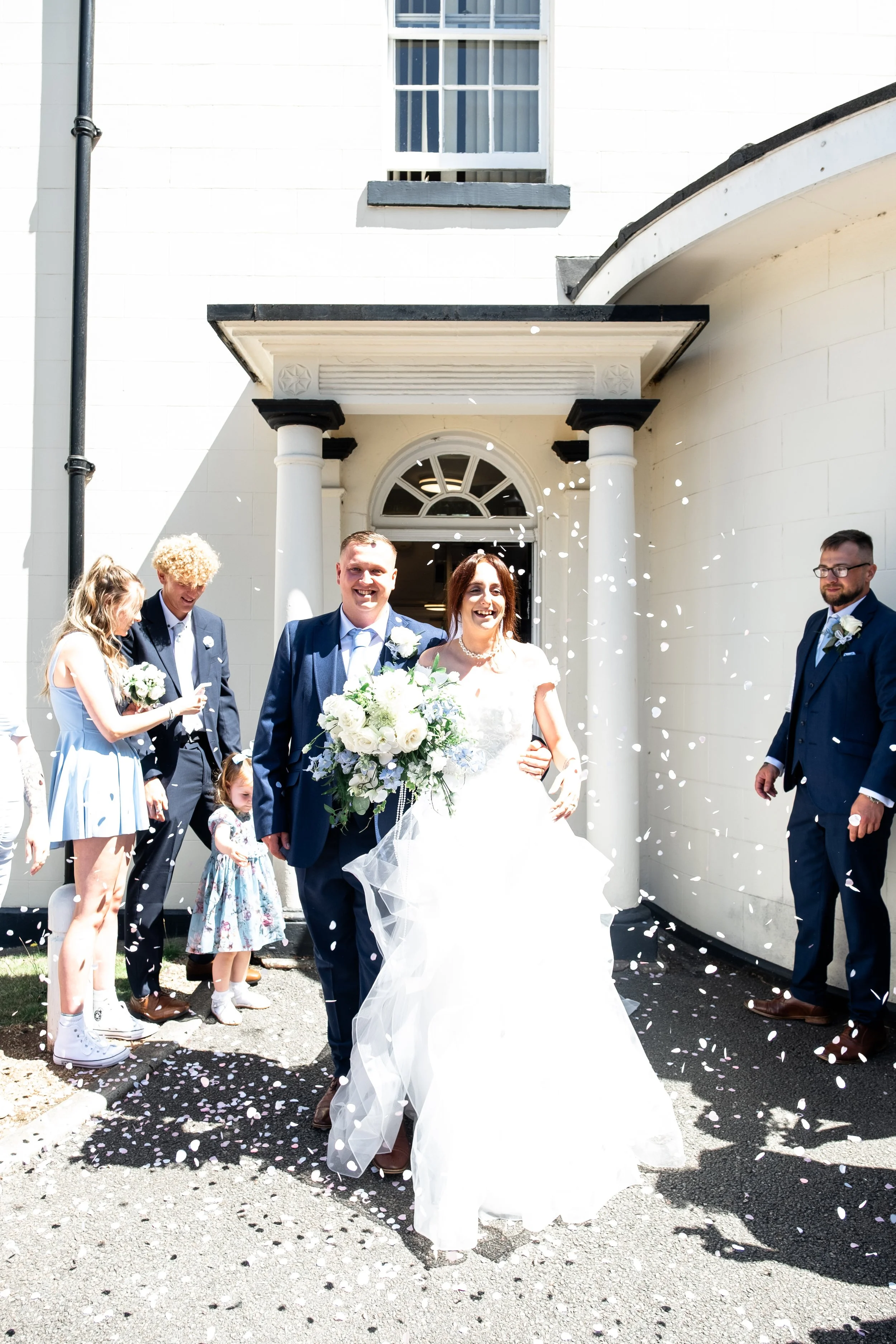 A bride and groom walking outside a white building, celebrating their wedding with guests throwing confetti.