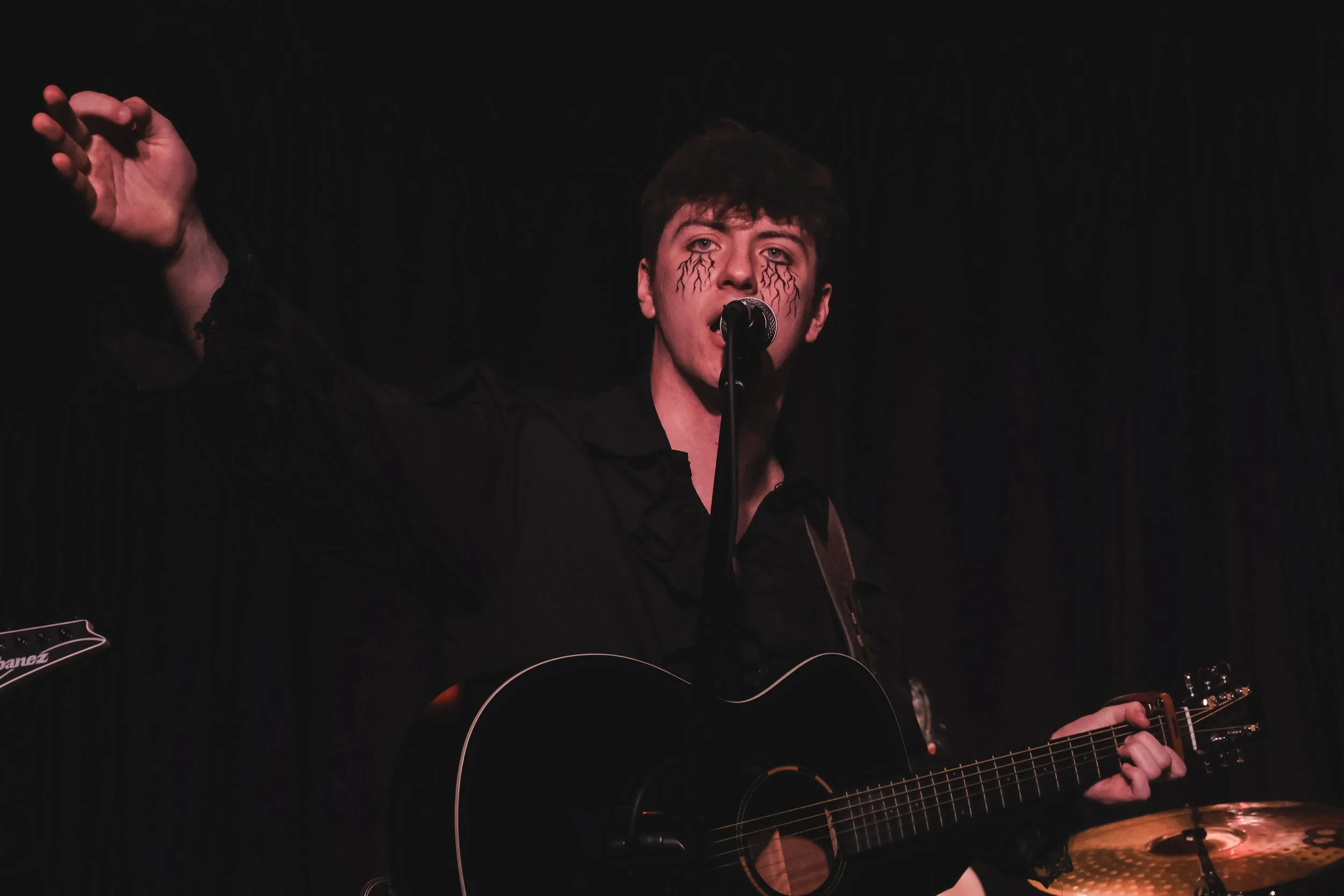 A young man with face paint resembling streams of lightening is singing into a microphone while playing an acoustic guitar on stage with a dark background.