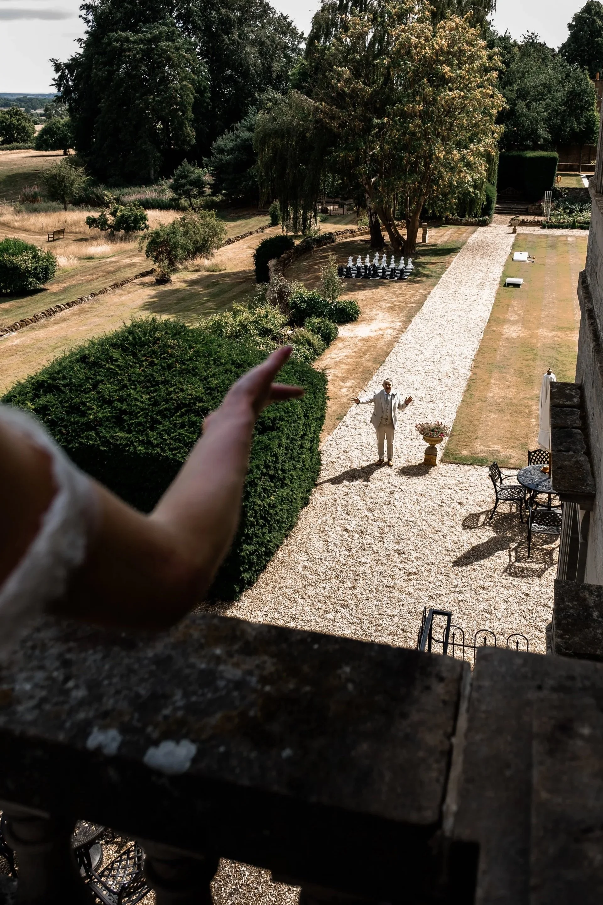 Person in the foreground pointing towards a man standing on a gravel path in a large, landscaped garden. The man has arms outstretched and is dressed in a white suit. The garden has structured pathways, bushes, trees, and outdoor furniture, with chess pieces visible in the background.