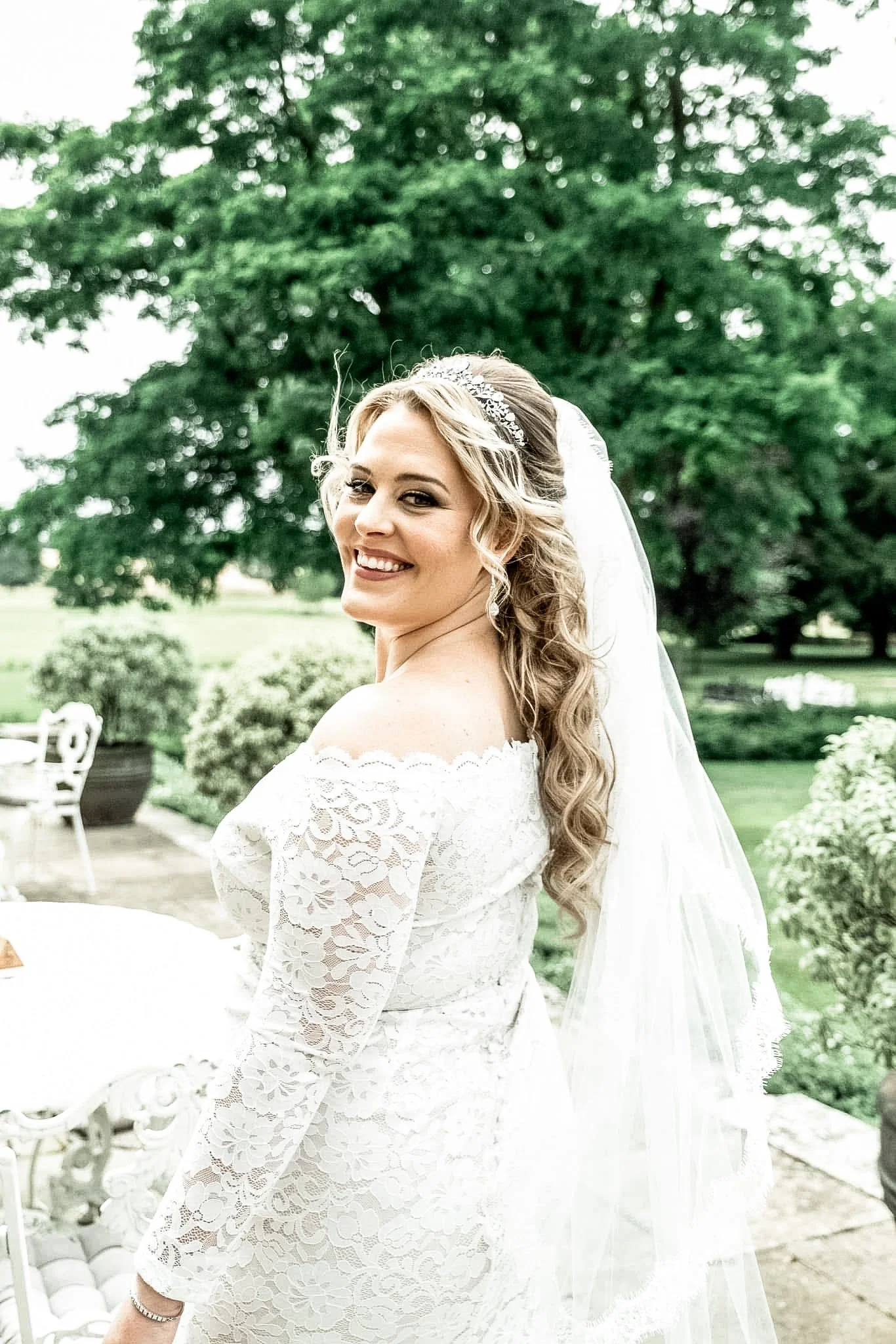 A bride in a white lace wedding dress with off-the-shoulder sleeves, smiling outdoors in front of large green trees, with a wedding veil, jewelry, and styled hair.