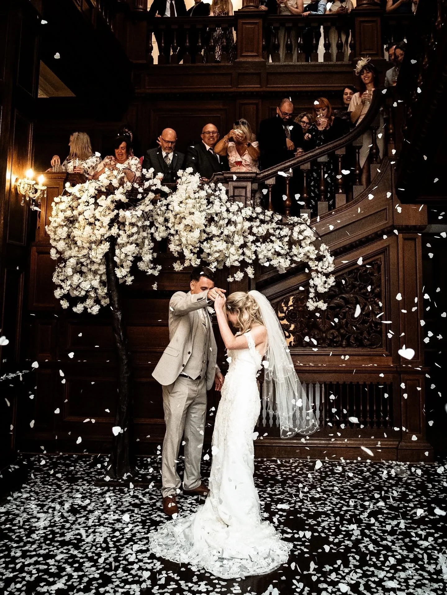 A bride and groom dancing at their wedding, with guests watching from a staircase above, surrounded by confetti and floral decorations.