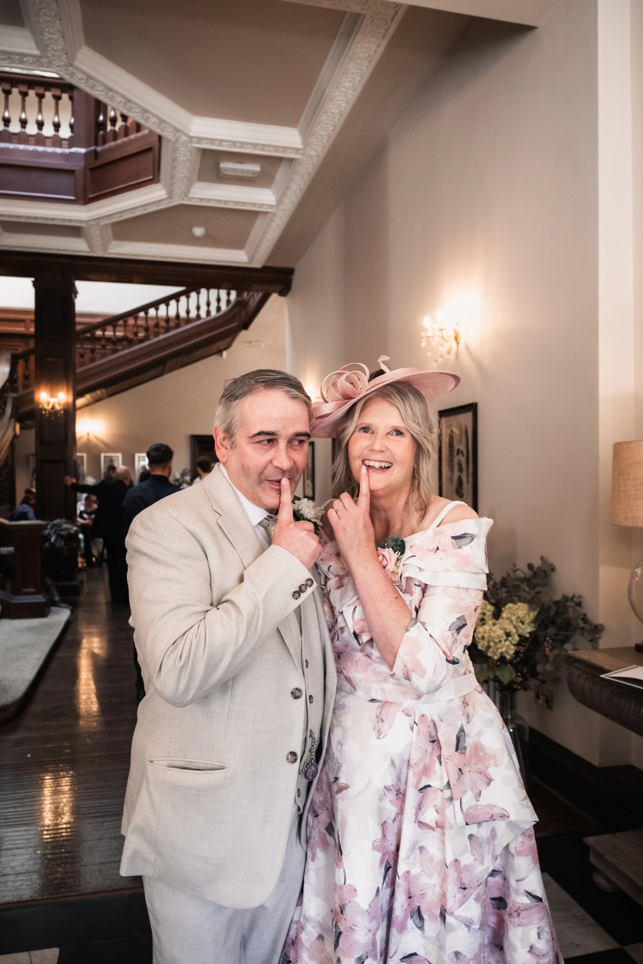 A man and woman at a wedding reception, both with their index fingers on their lips, making a shh gesture. The woman is wearing a floral dress and a pink hat. The man is dressed in a light-colored suit.