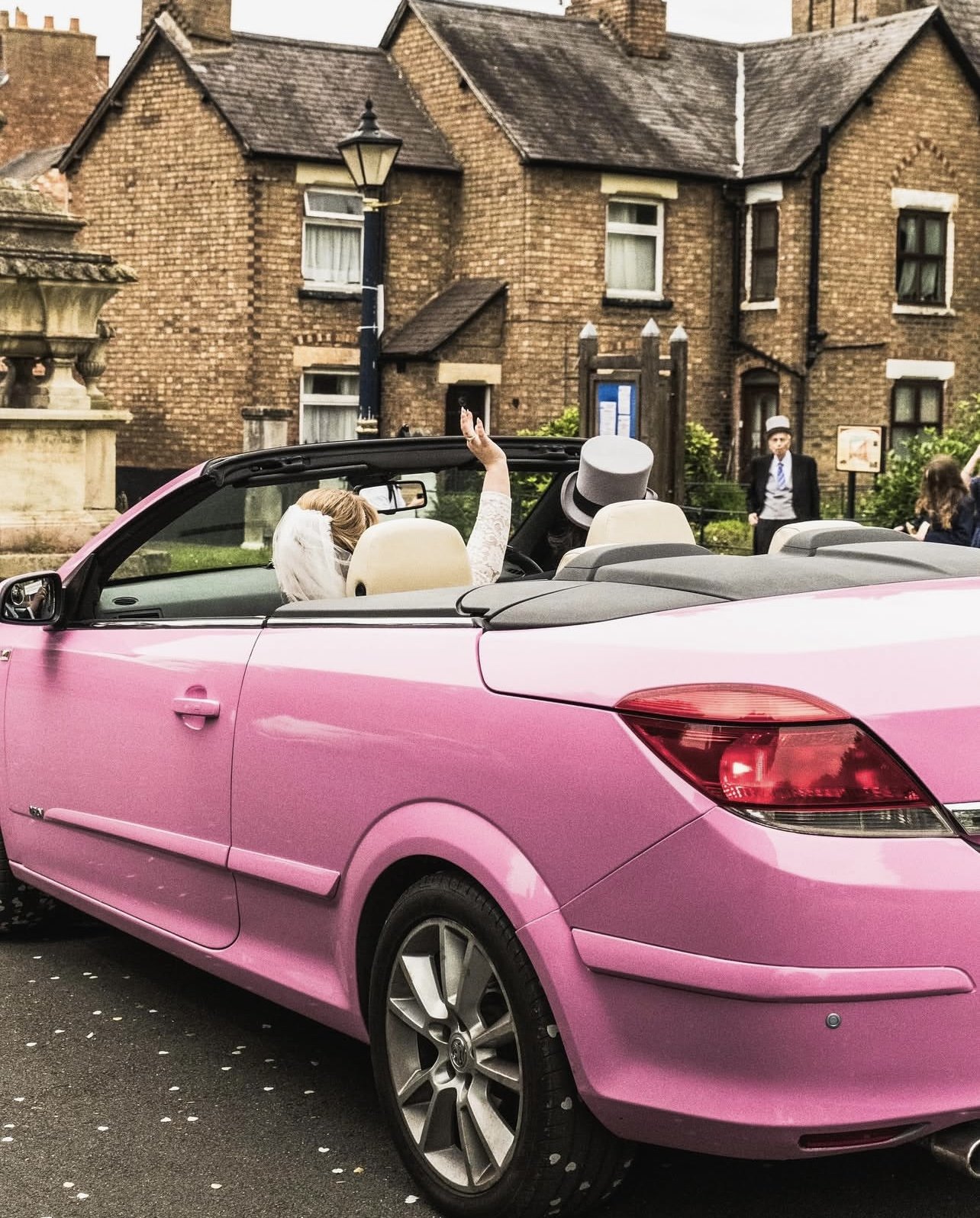 A pink convertible car with a bride and a groom inside, the bride waving and wearing a veil and the groom wearing a gray hat. There are people in the background, some dressed formally, and an old brick building behind them.