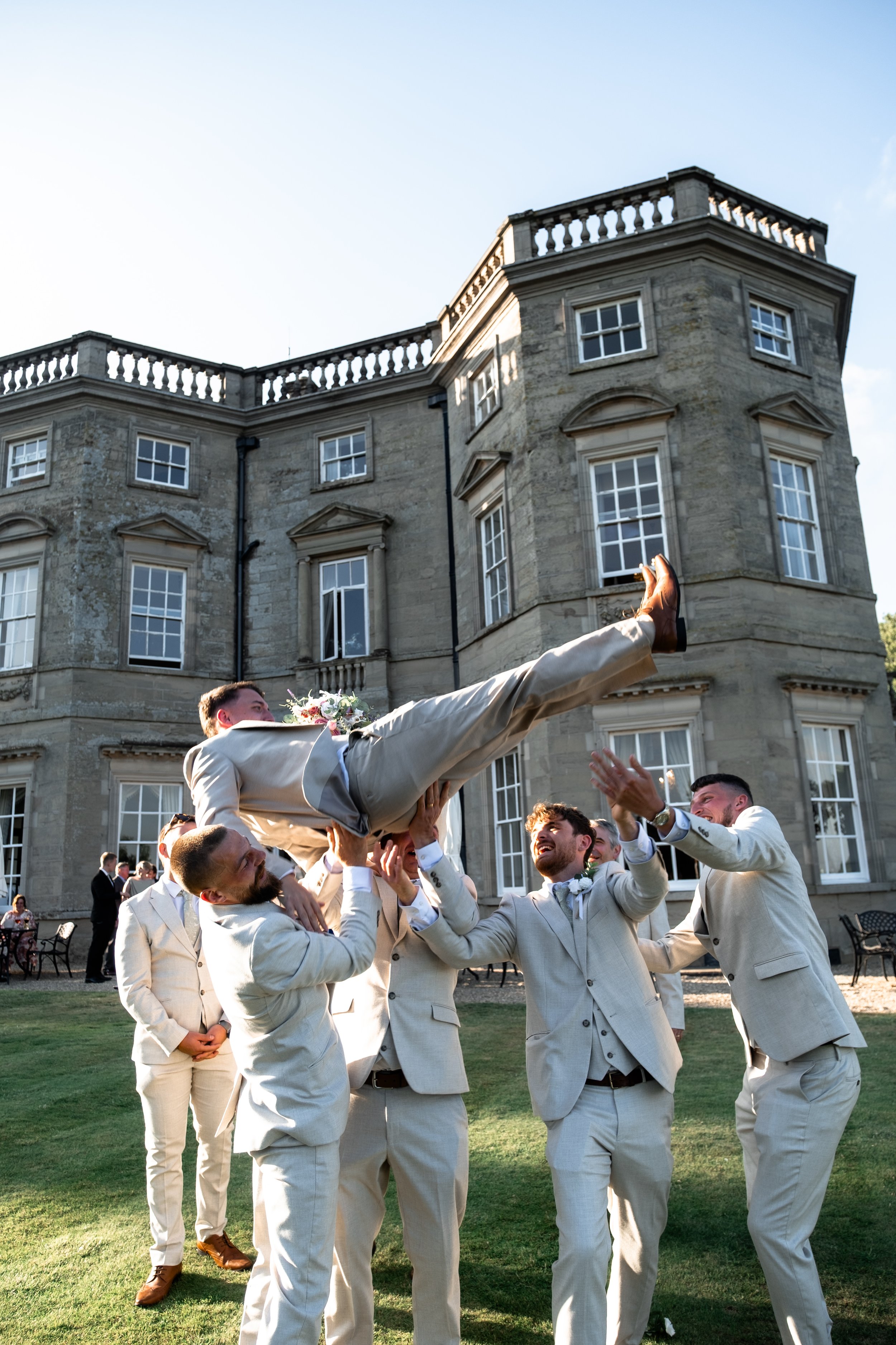 Groom being lifted in the air by groomsmen outside a large stone mansion during a wedding celebration.