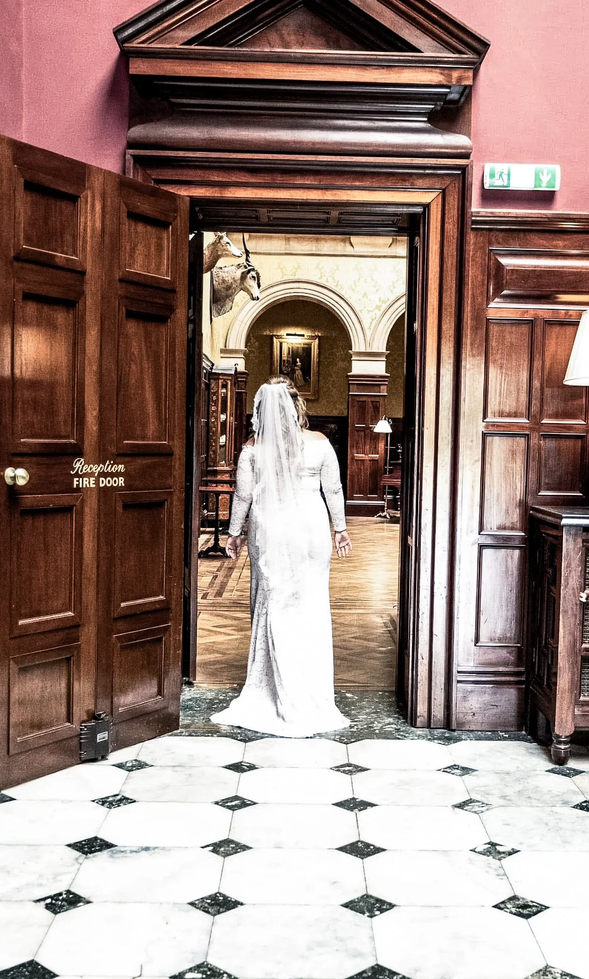 A bride in a long white dress and veil walking through a wooden doorway into a room with ornate woodwork, paintings, and mounted animal heads, with black and white patterned tile flooring.