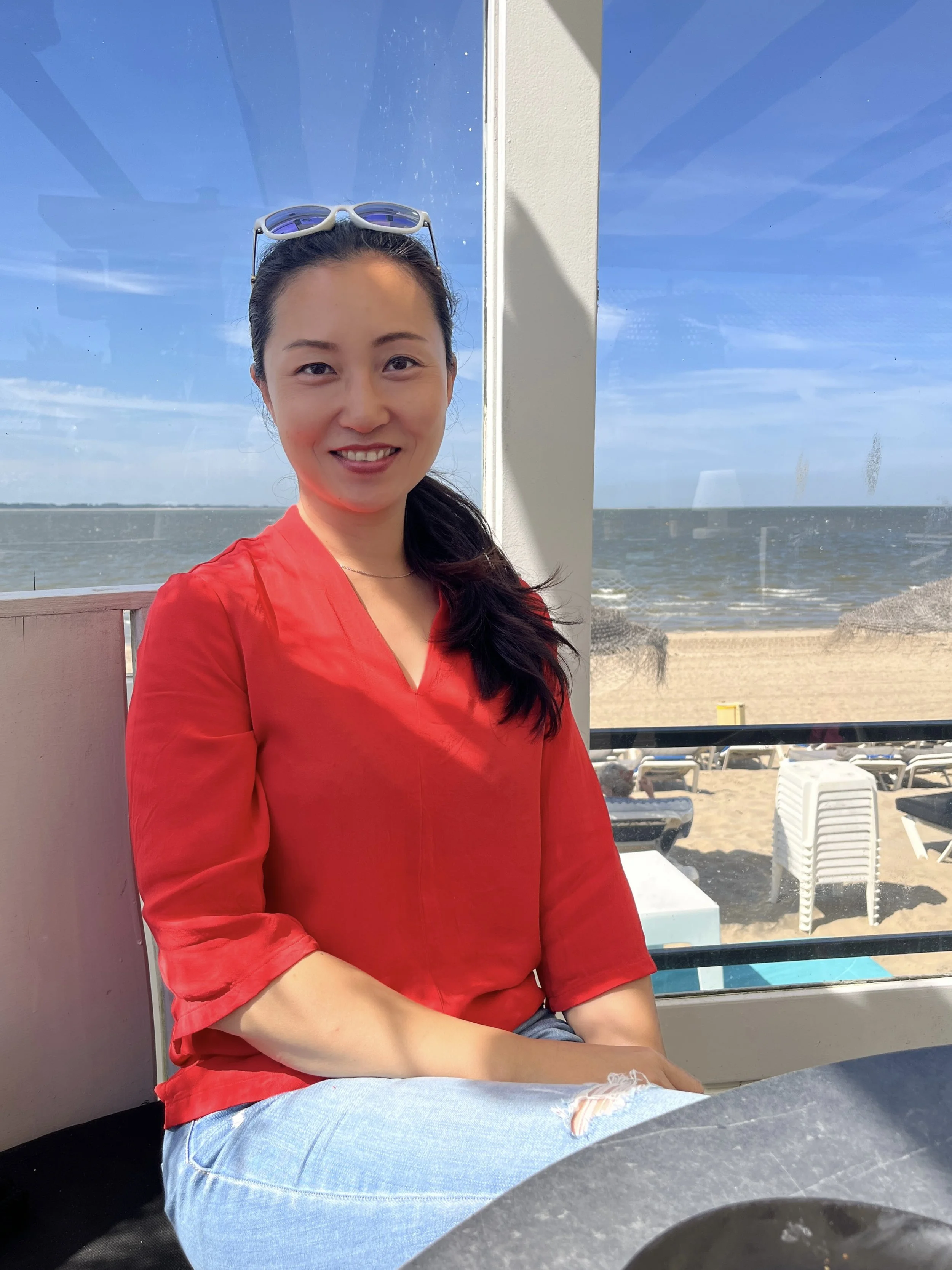 Young woman with dark hair, wearing sunglasses on her head and a red shirt, sitting indoors with a window behind her showing a beach and ocean scene.
