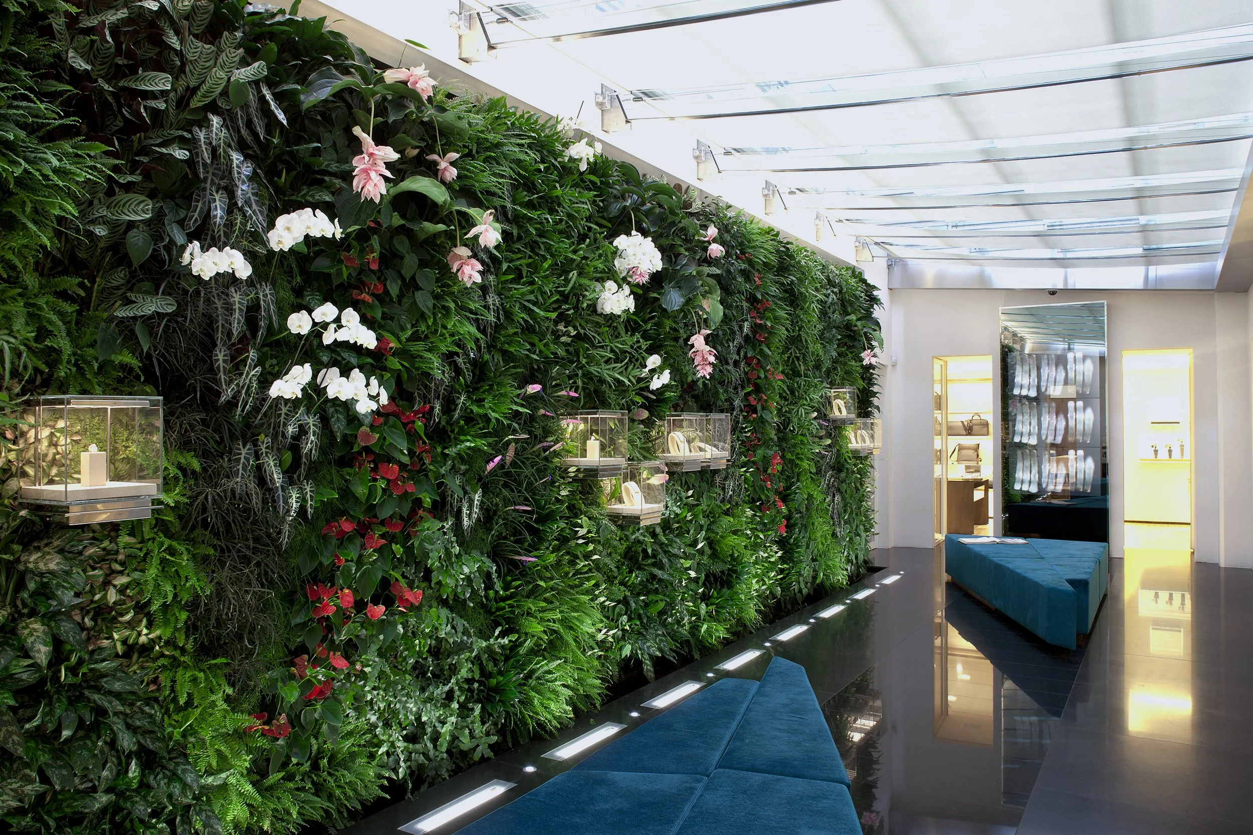 Interno di un negozio con parete verde di piante e fiori, illuminate da luci a soffitto, con sedie blu e vetrine espositive.
