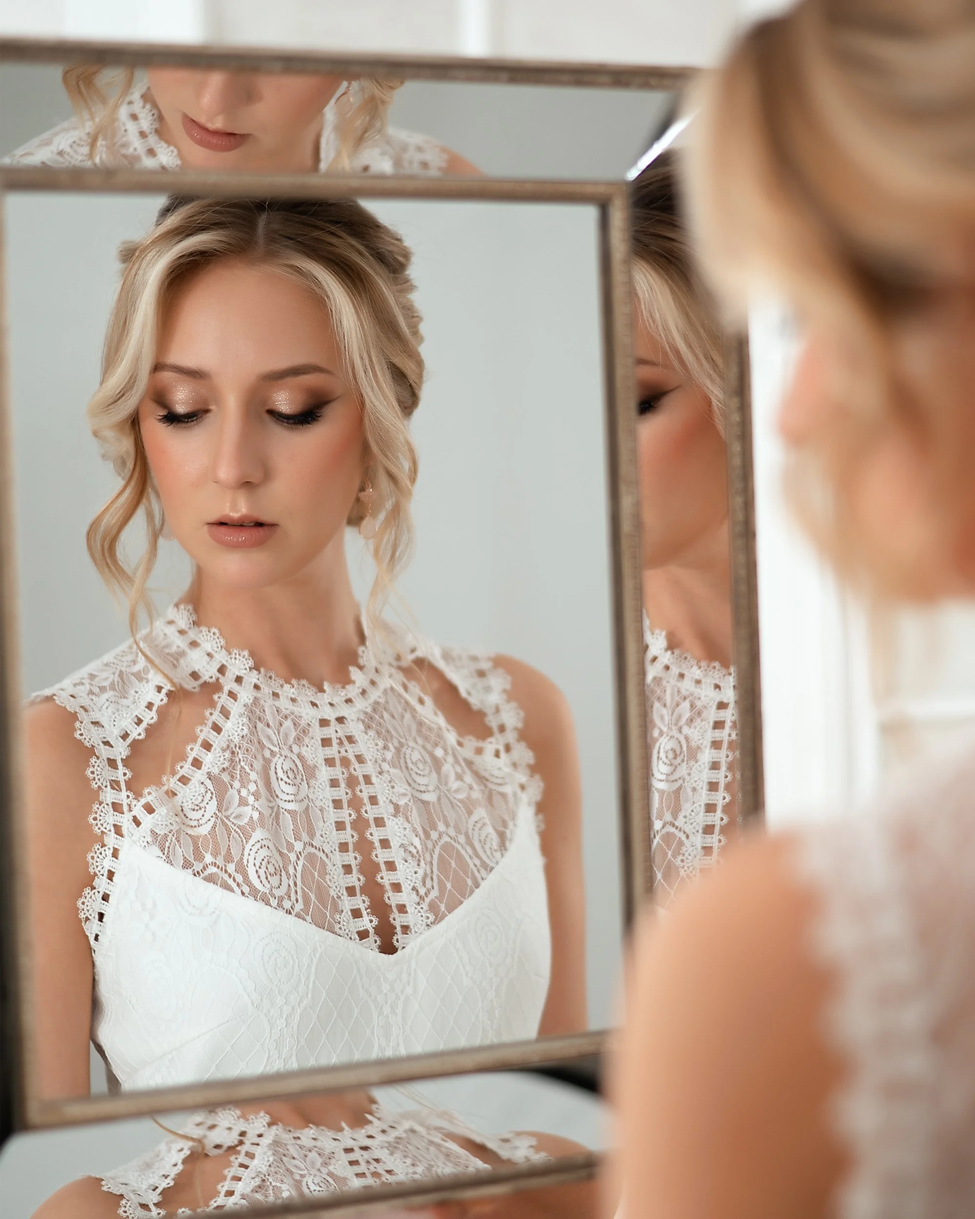 Junge Frau in weißem Spitzenkleid betrachtet sich in einem Spiegel.