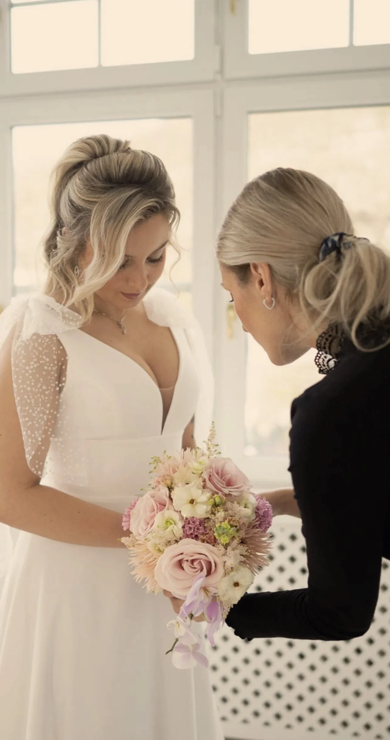 Zwei Frauen, eine in Hochzeitskleid und die andere in dunklem Kleid, bei einer Hochzeitszeremonie, die ein Brautstrauß hält.
