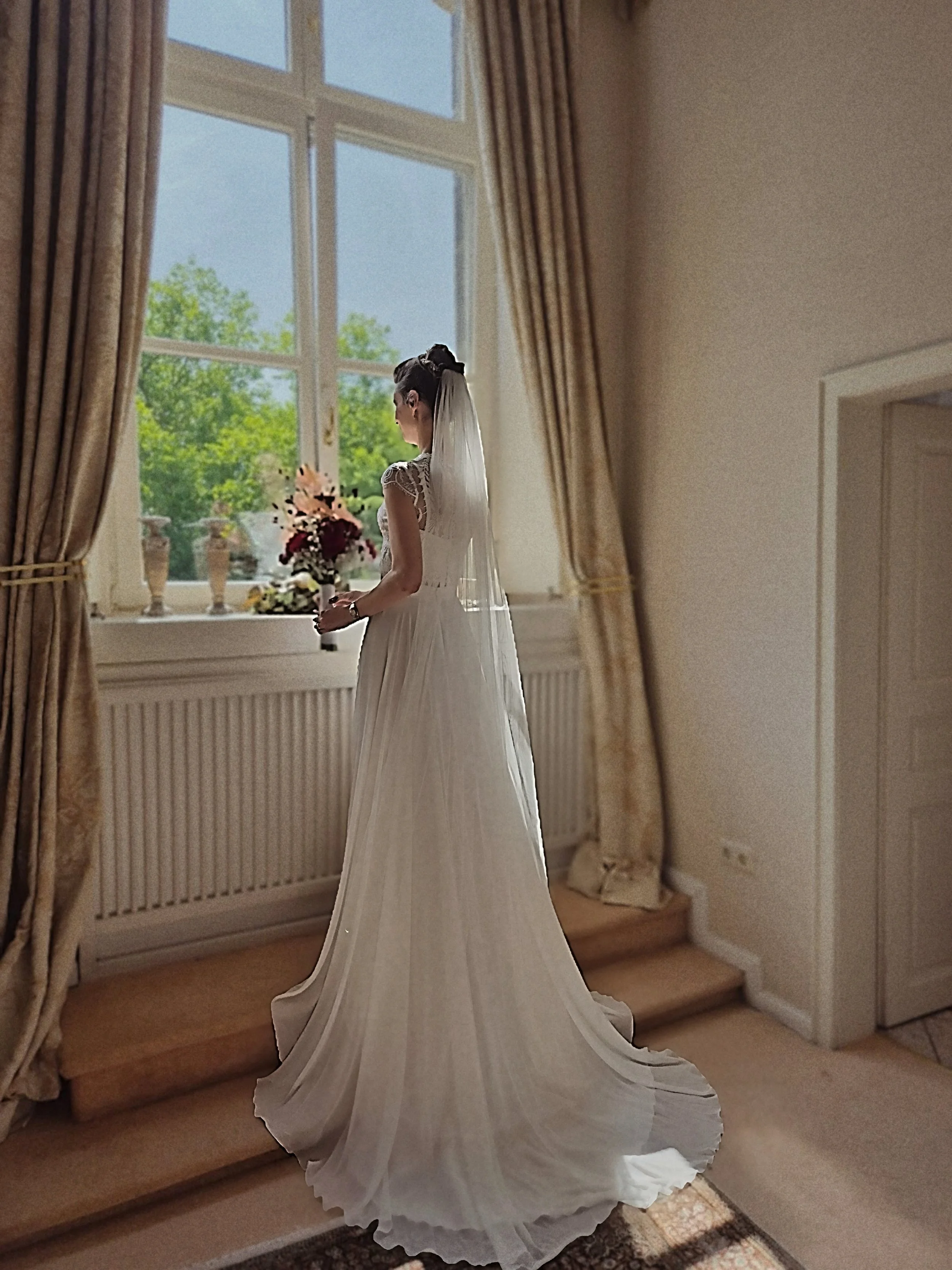 Eine Braut in einem weißen Brautkleid mit langem, plissiertem Rock steht vor einem großen Fenster, hält einen Blumenstrauß und blickt nach draußen.