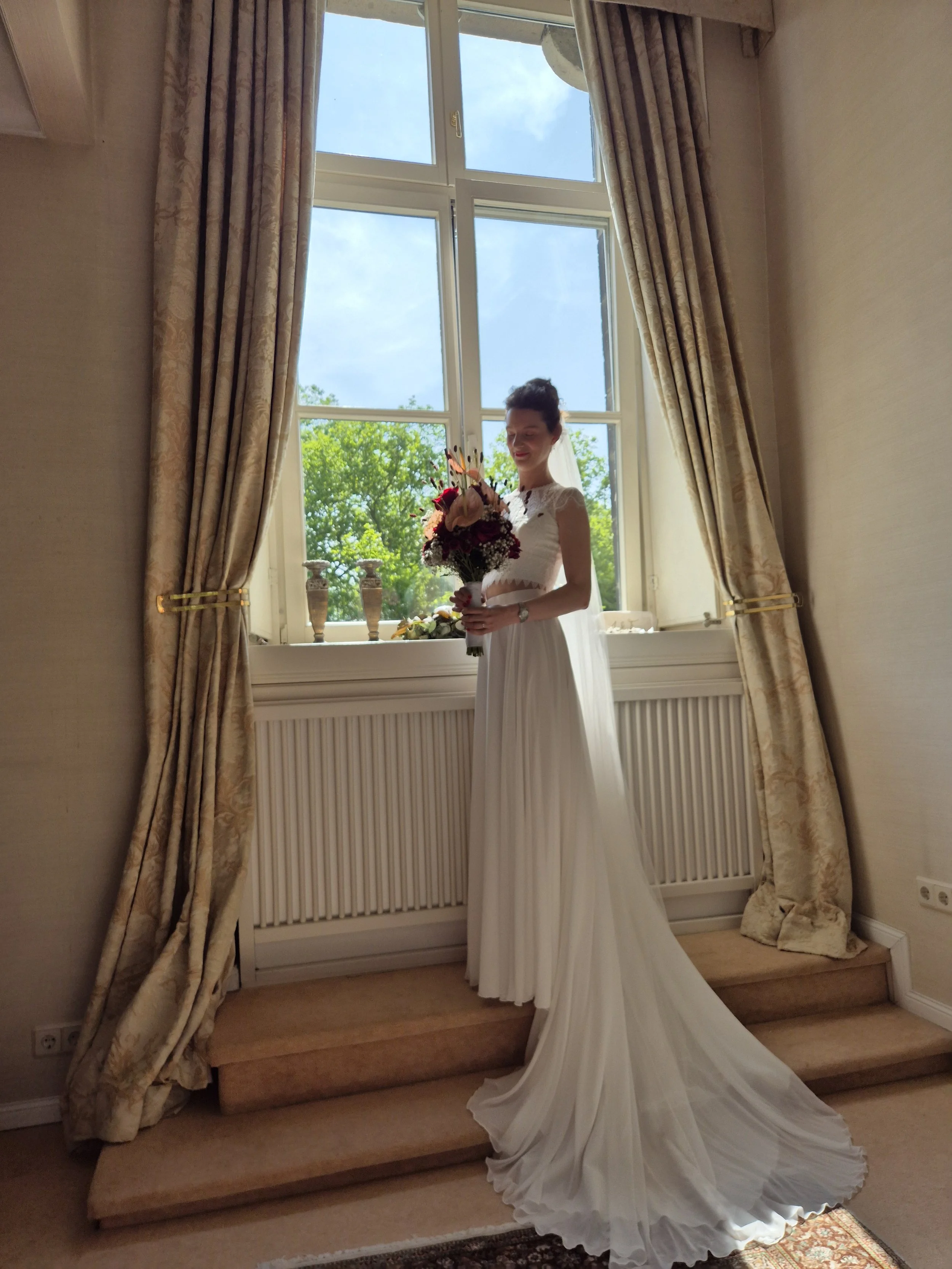 Braut in weißem Brautkleid steht vor einem großen Fenster, hält einen Blumenstrauß und lächelt, hinter ihr blauer Himmel und grüne Bäume.