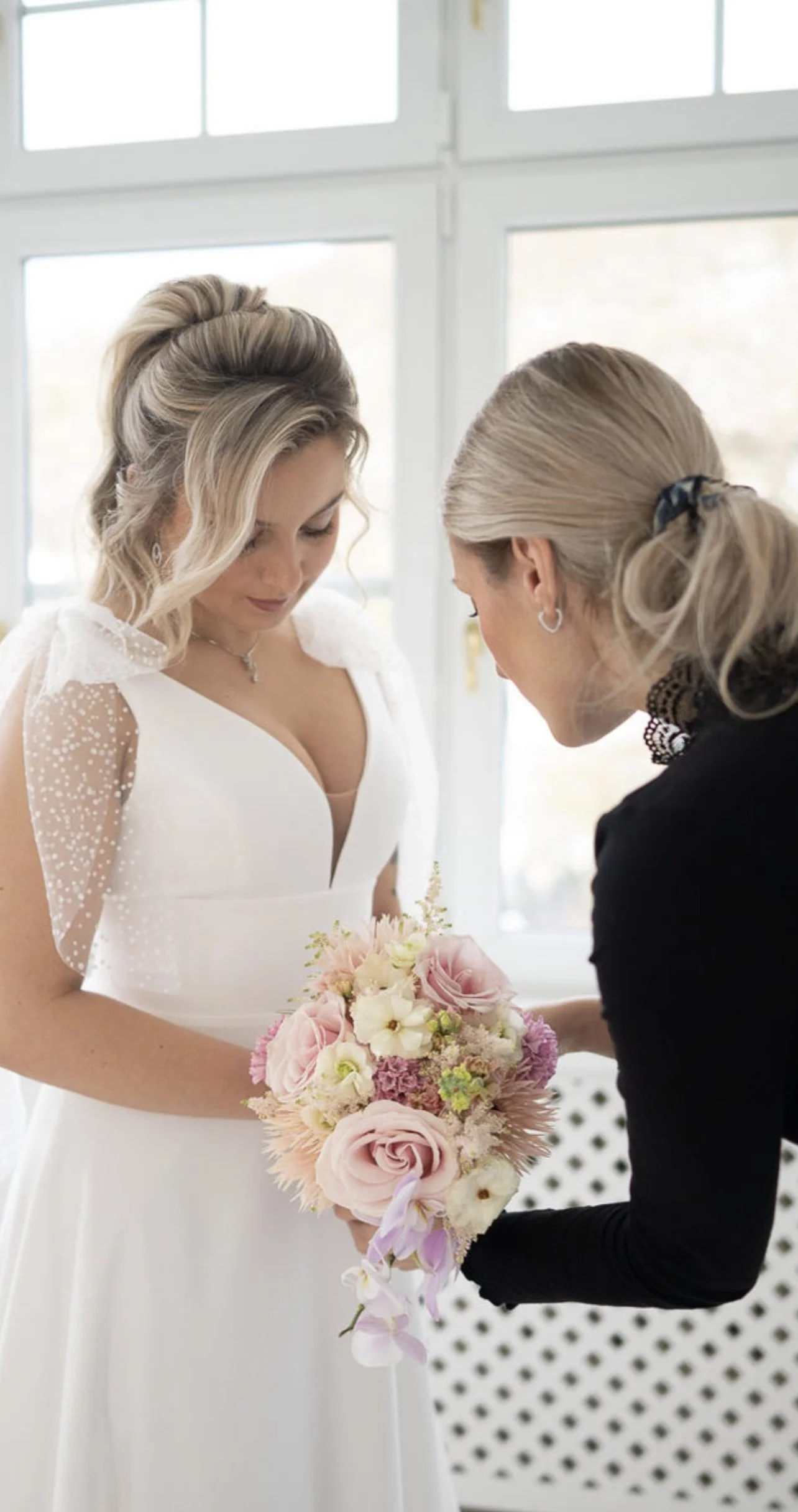 Eine Braut in einem weißen Kleid hält einen Blumenstrauß, während eine Frau im schwarzen Kleid ihr eine Blume überreicht.