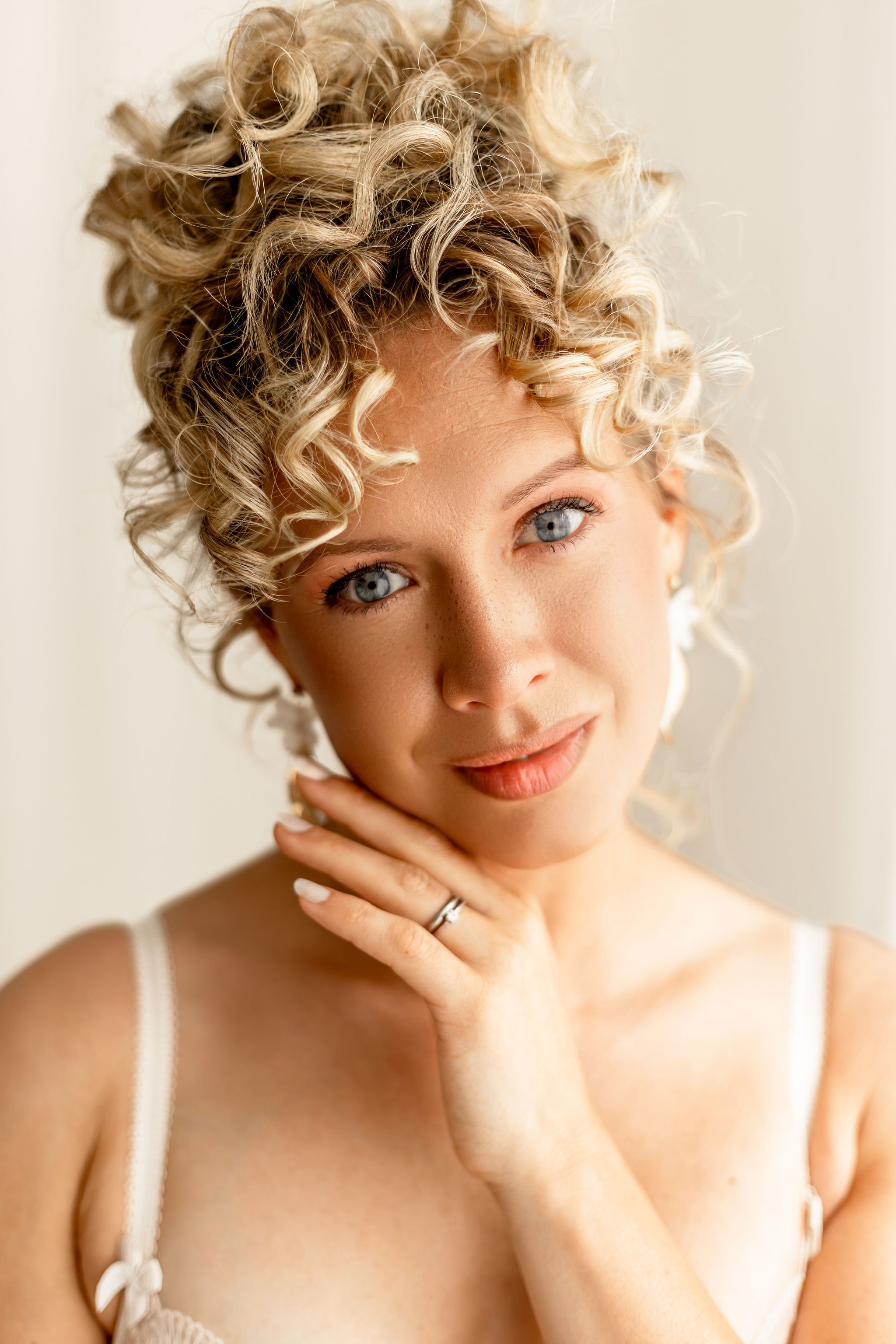 Nahaufnahme eines lächelnden jungen Mädchens mit blauen Augen und lockigem blonde Haar, das ihre Hand mit einem Ring am Finger an ihrem Kinn hält.