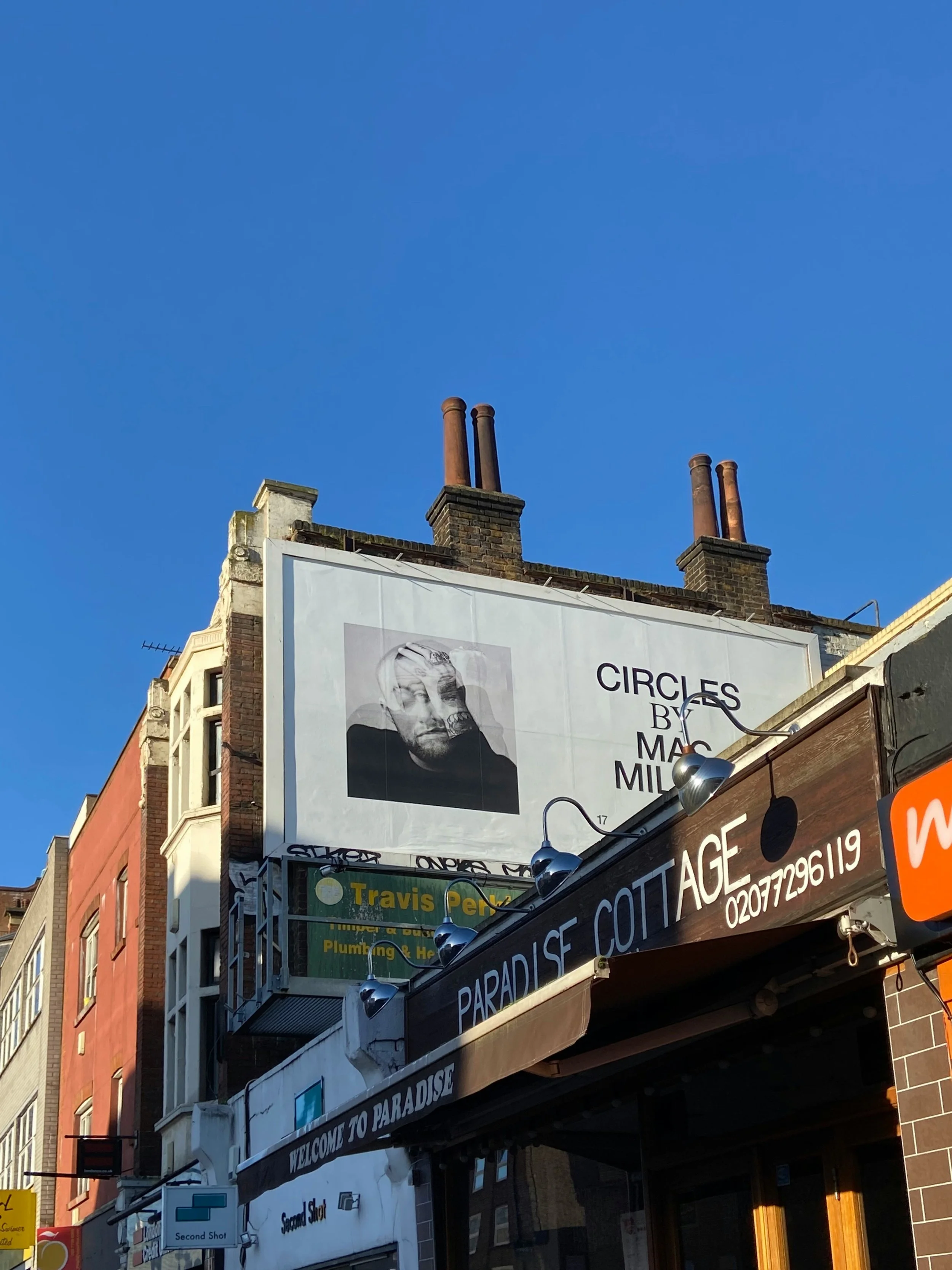City buildings with a large billboard advertising an art exhibit titled 'Circles' by Mac Miller. The billboard features an abstract portrait of a man's face. The sky is clear and blue.