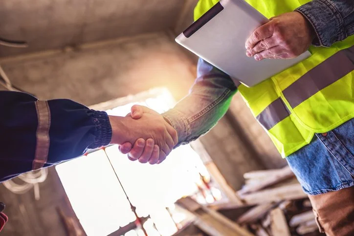 Two people shaking hands, one wearing a high-visibility safety vest and the other in a dark jacket, inside a barn or construction site, with one holding a clipboard.