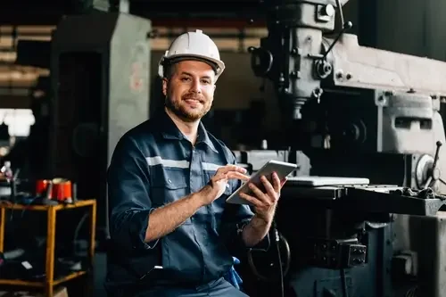A man wearing a white safety helmet and dark work clothes, smiling and using a tablet in an industrial workshop with machinery in the background.