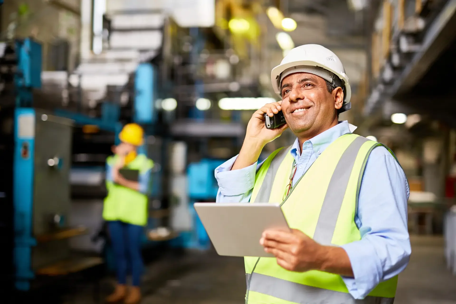 A smiling male industrial worker talking on a cell phone while holding a tablet in a factory setting, with another worker in the background.
