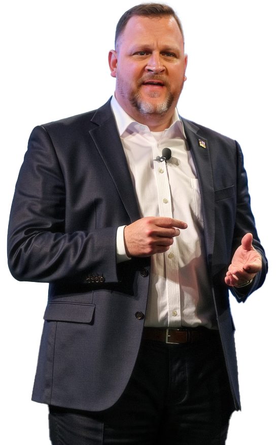 Man with short hair and a beard, wearing a dark suit and white shirt, giving a speech or presentation with a microphone clipped to his shirt, against a background with red, blue, and white colors.