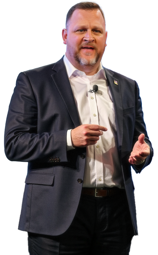 Man with short hair and a beard, wearing a dark suit and white shirt, giving a speech or presentation with a microphone clipped to his shirt, against a background with red, blue, and white colors.